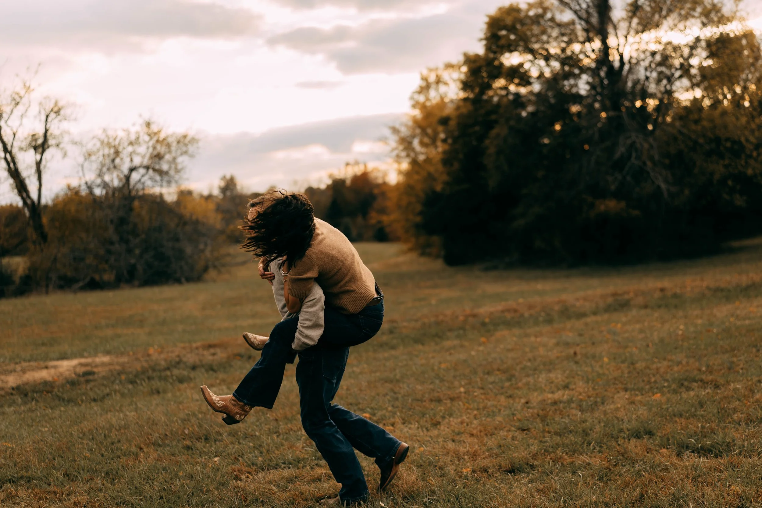 A couple playing and spinning in a grassy field during sunset, surrounded by fall trees.