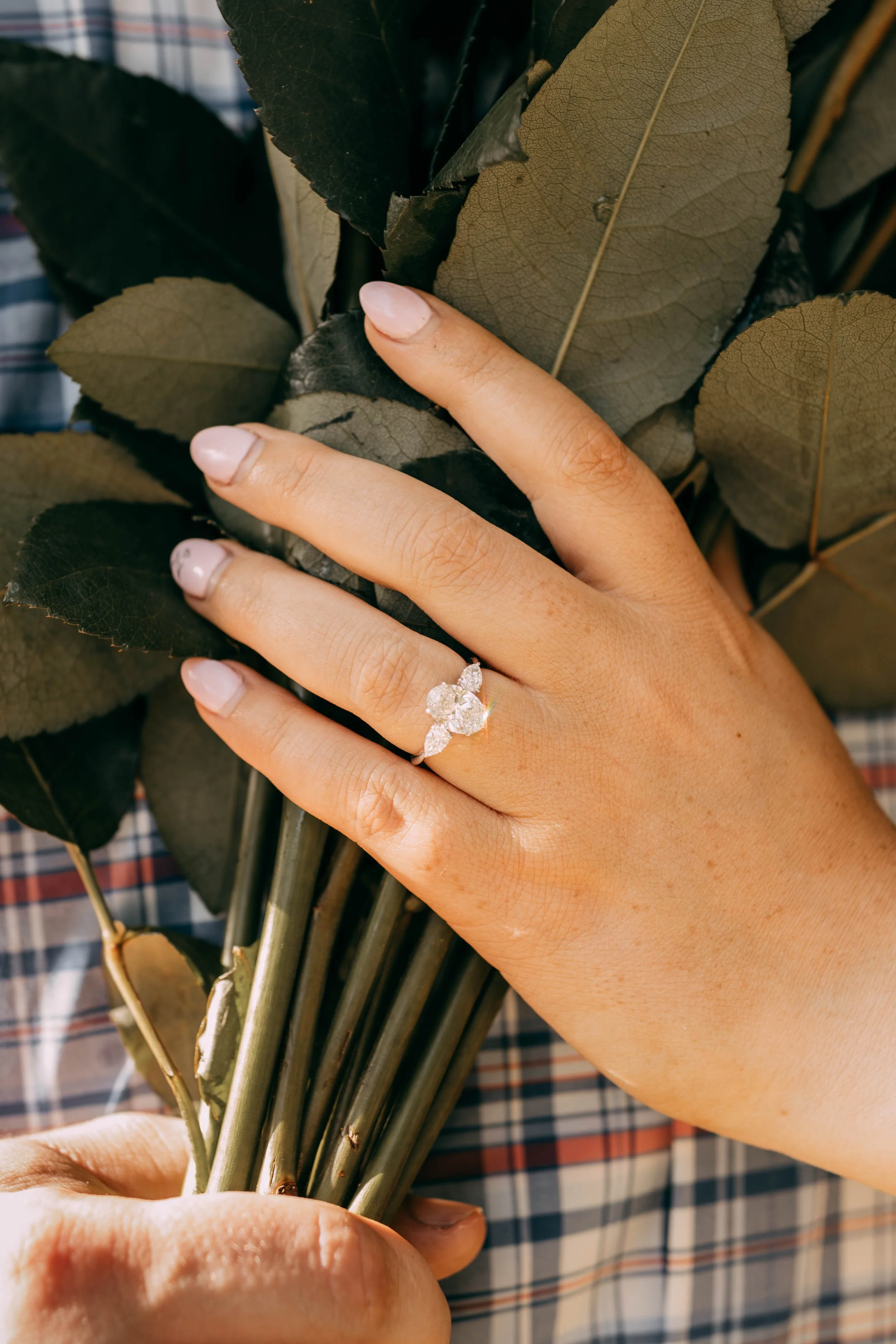 A person's hand with a diamond engagement ring on the ring finger, holding a bunch of dark green and brown leaves. The nails are painted in a pale pink color. The background is a plaid fabric.