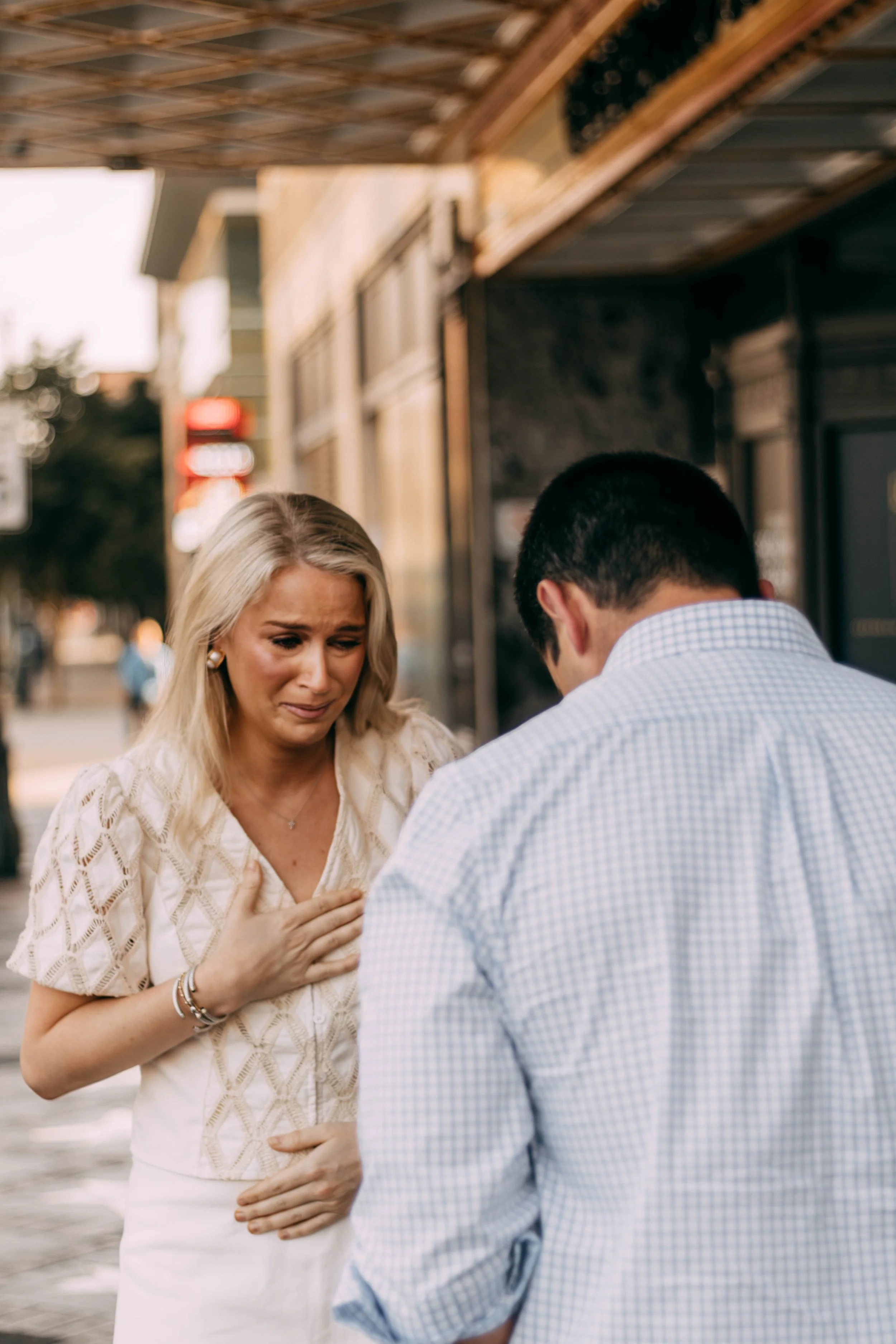 A woman appears emotional, clutching her chest and stomach, as she prays or begs in front of a man, outside on a sidewalk.