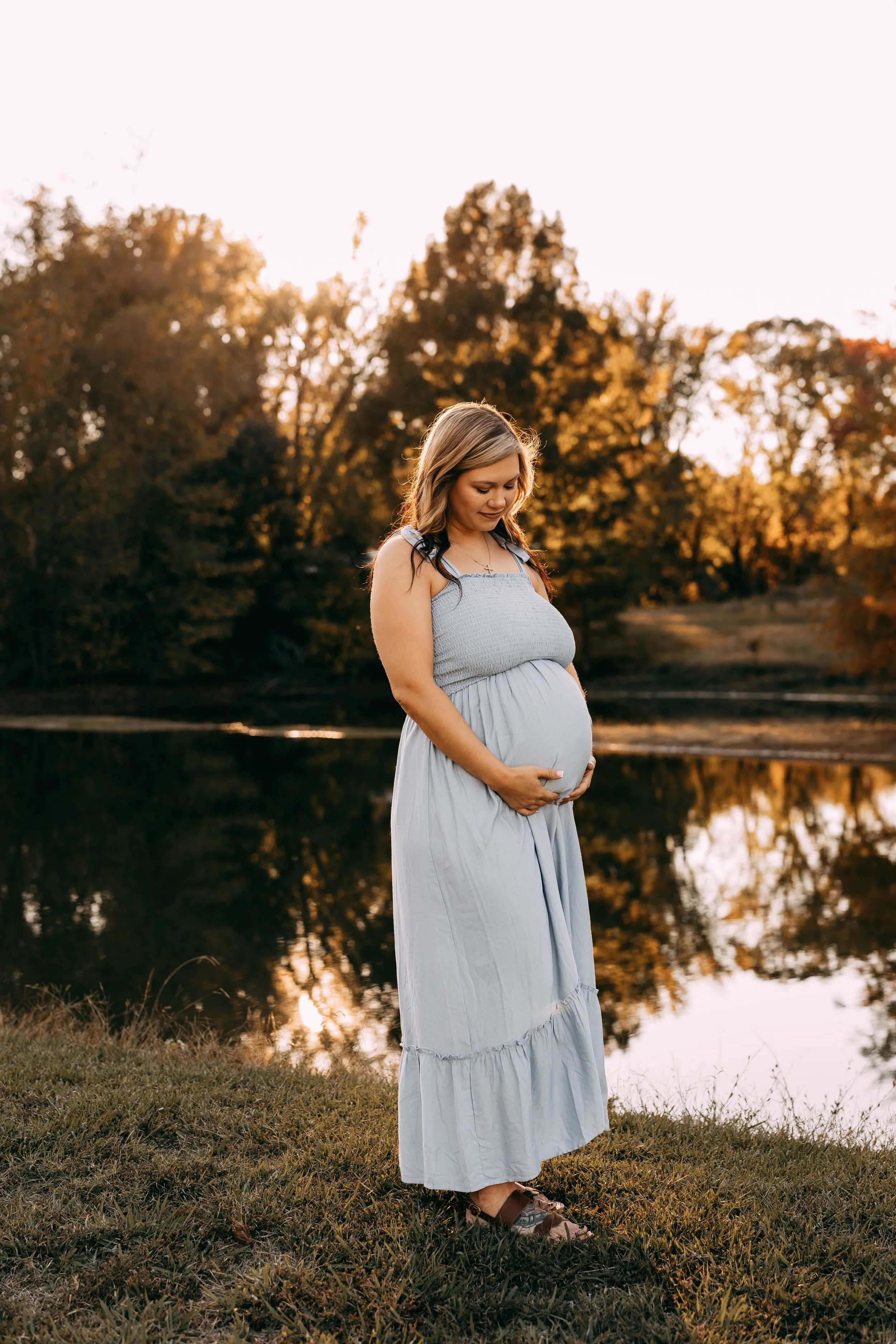 A pregnant woman in a light blue dress standing by a lake during sunset, holding her belly and looking down, with trees in the background.