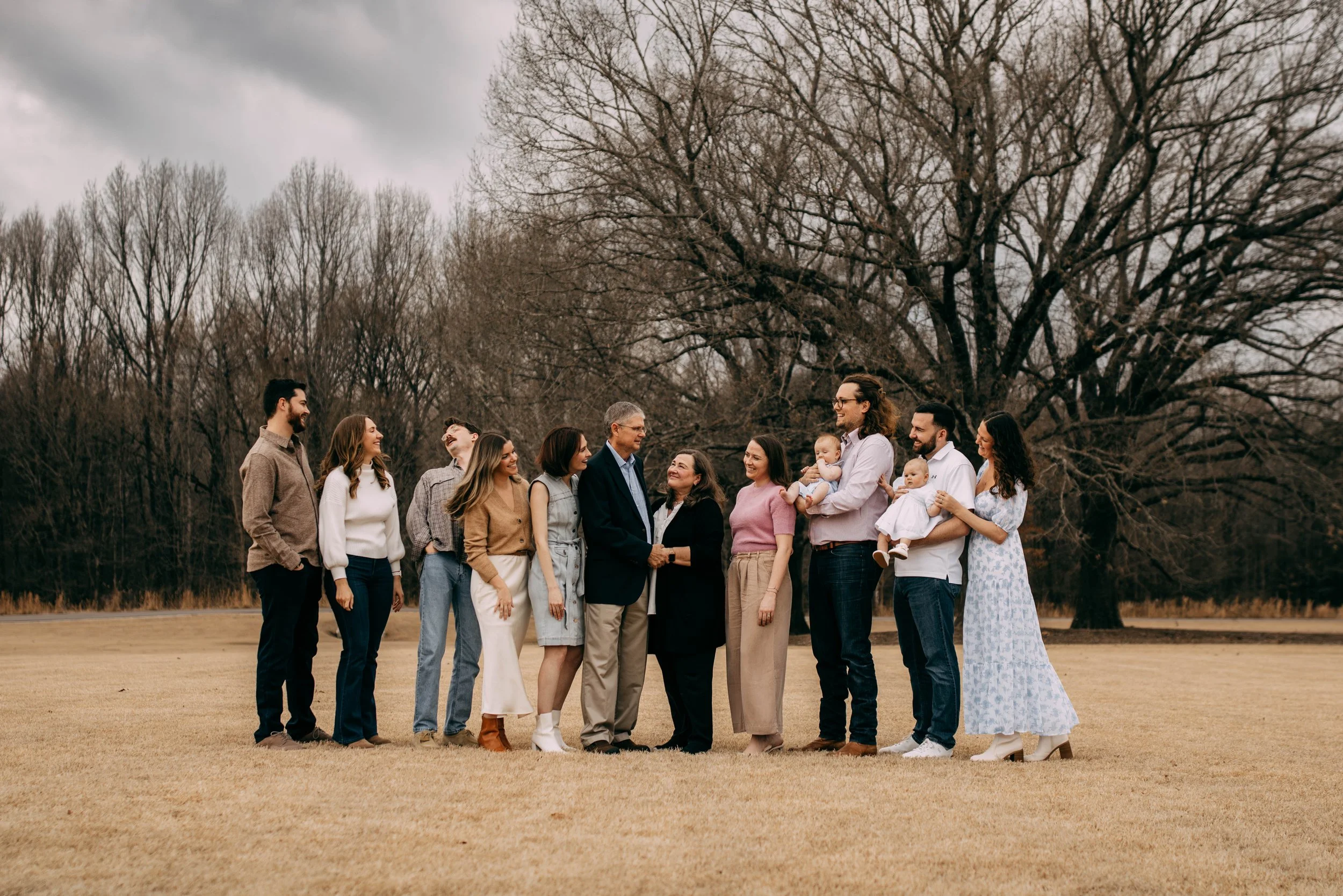 A large family gathering outdoors featuring multiple generations, including elderly, adults, children, and babies, standing on a grassy field with a large leafless tree in the background on a cloudy day.