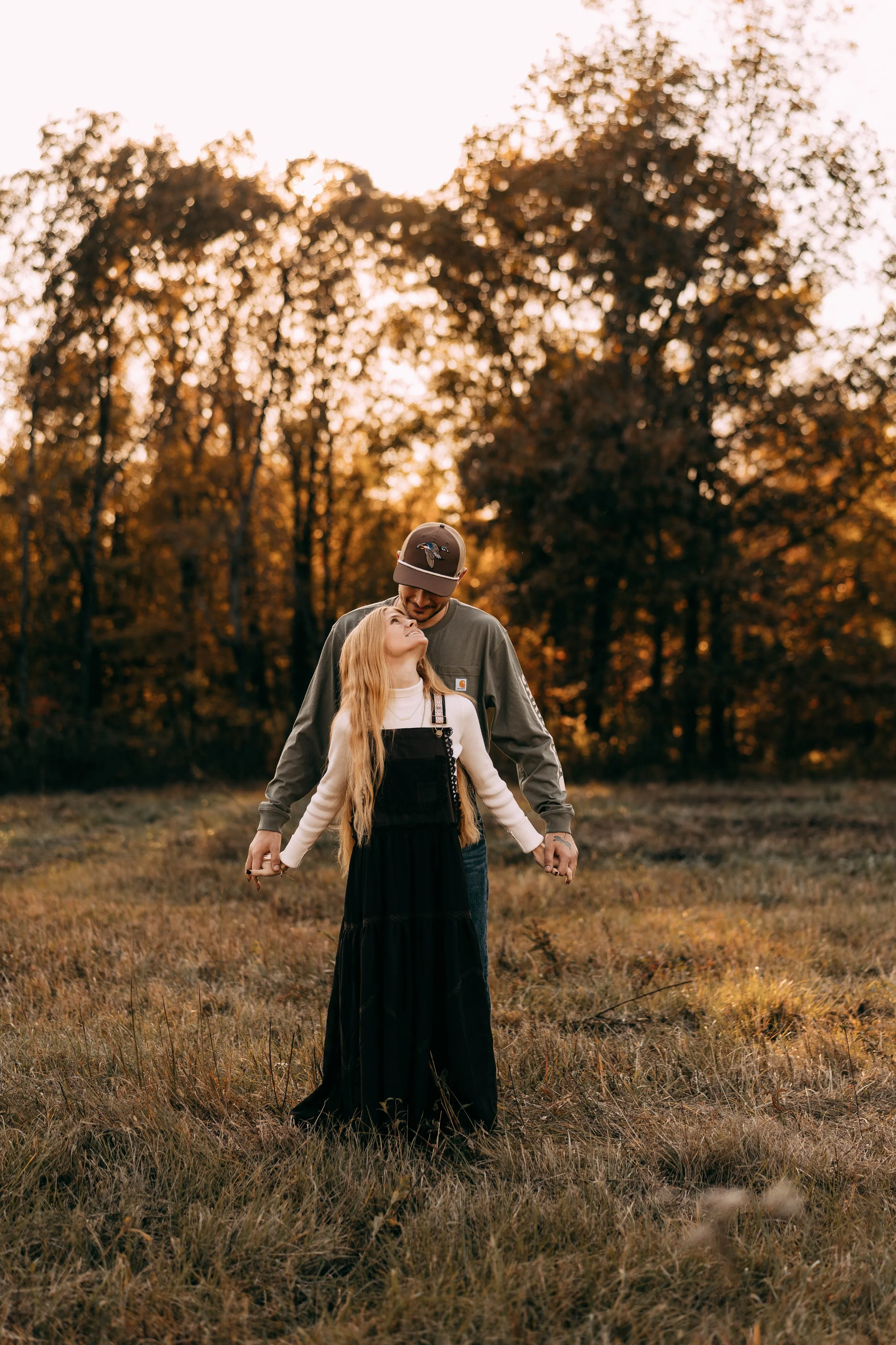 A man and a young girl standing close together in a field with trees in the background during sunset, holding hands and sharing a happy moment.