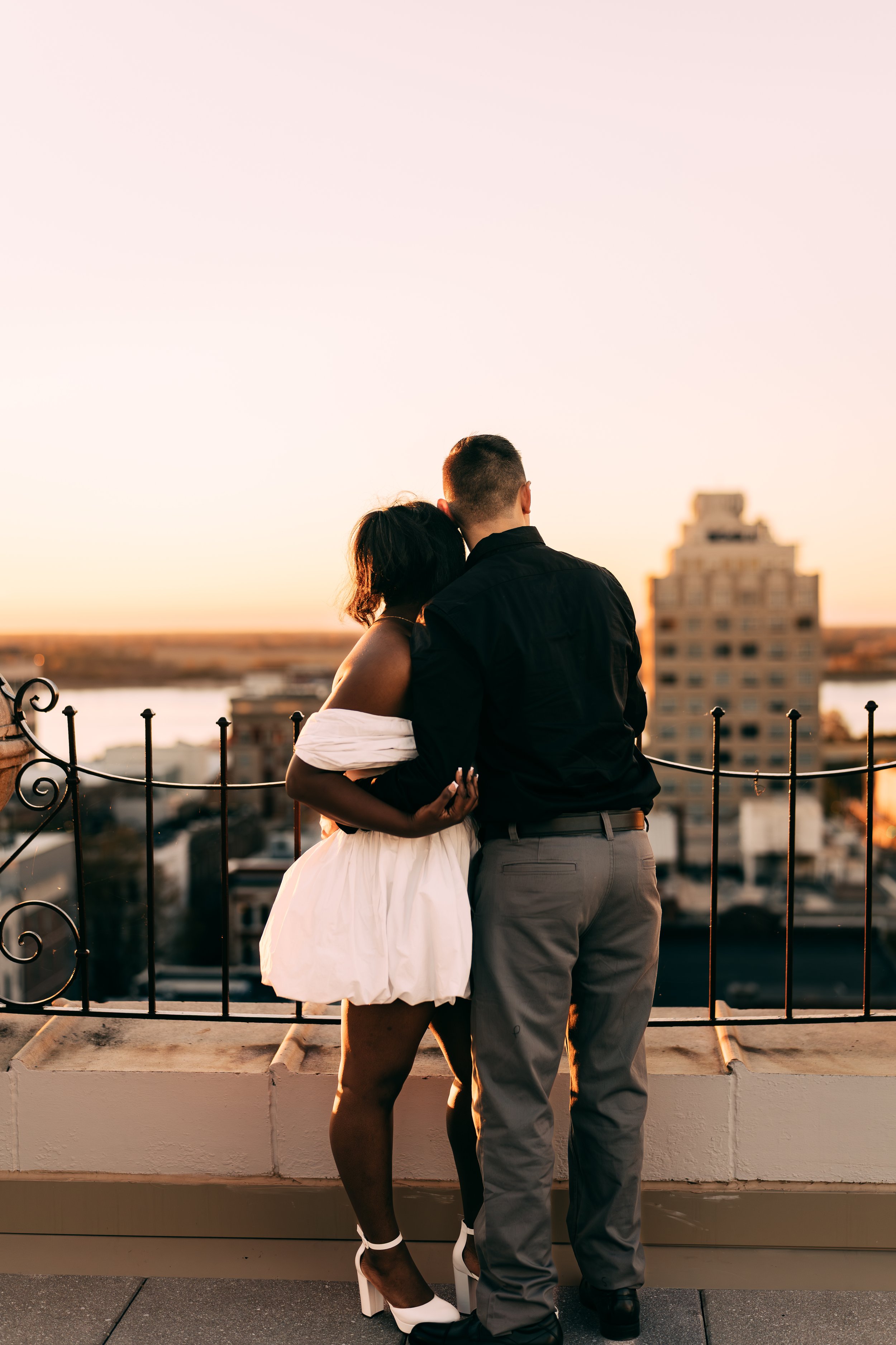 A couple stands close together on a rooftop at sunset, with the city skyline and river in the background.