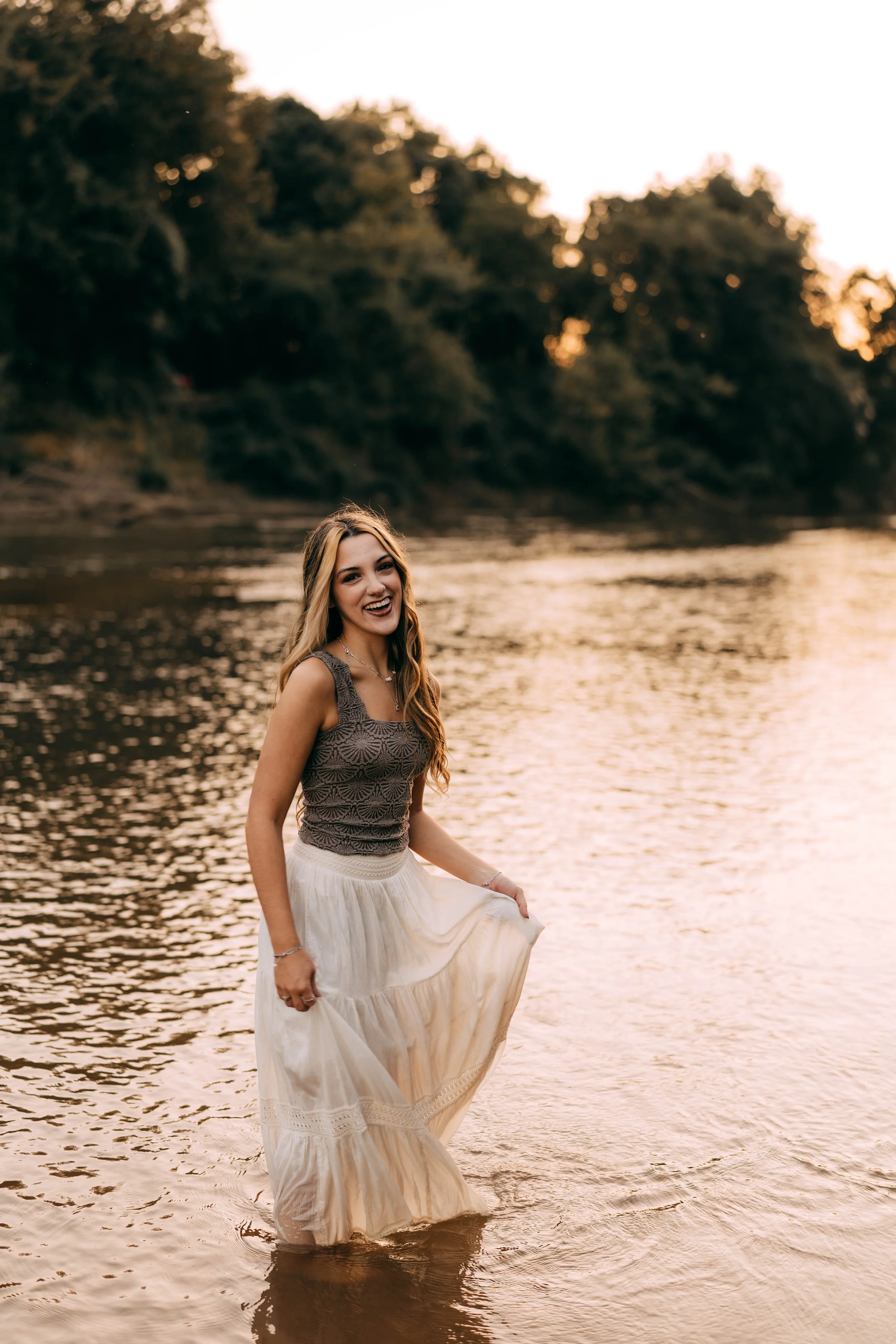 A young woman with long, wavy blonde hair, smiling, standing in a river at sunset, wearing a sleeveless brown top and a long white skirt.