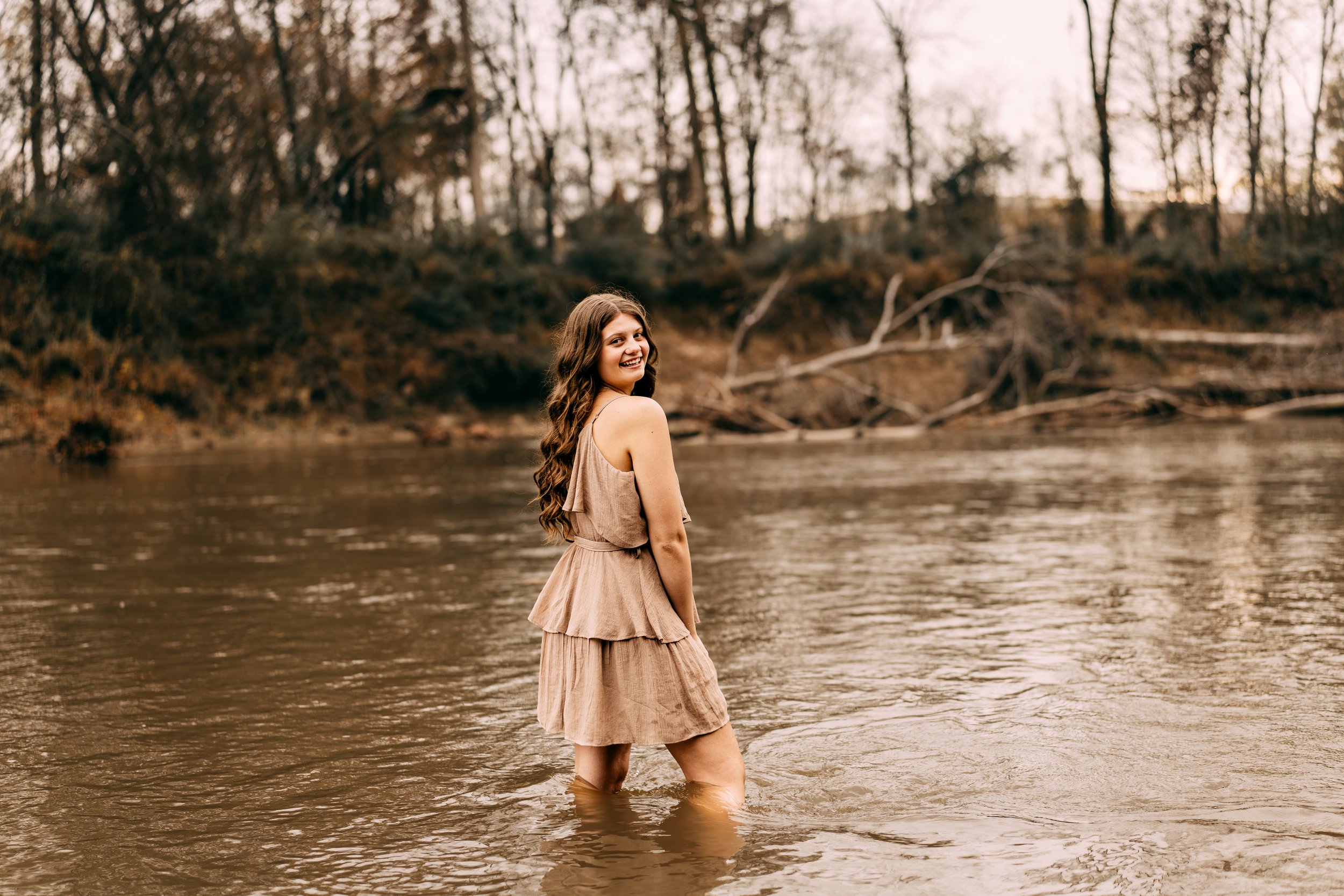 A woman with long brown hair wearing a sleeveless beige dress stands in a river, smiling and looking back at the camera, with trees and fallen branches in the background.