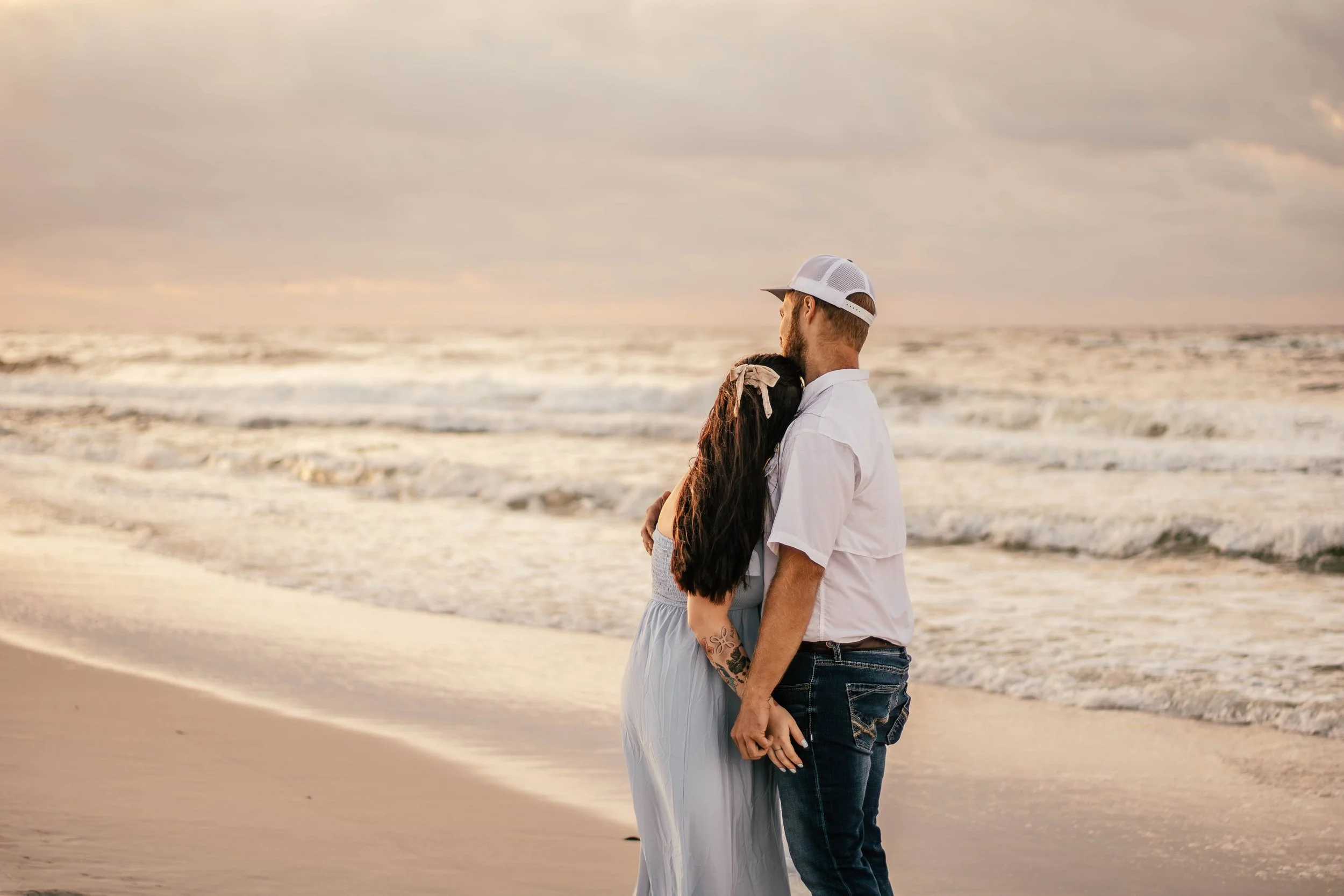 A couple standing on the beach at sunset, with the woman hugging the man and both looking out at the ocean.