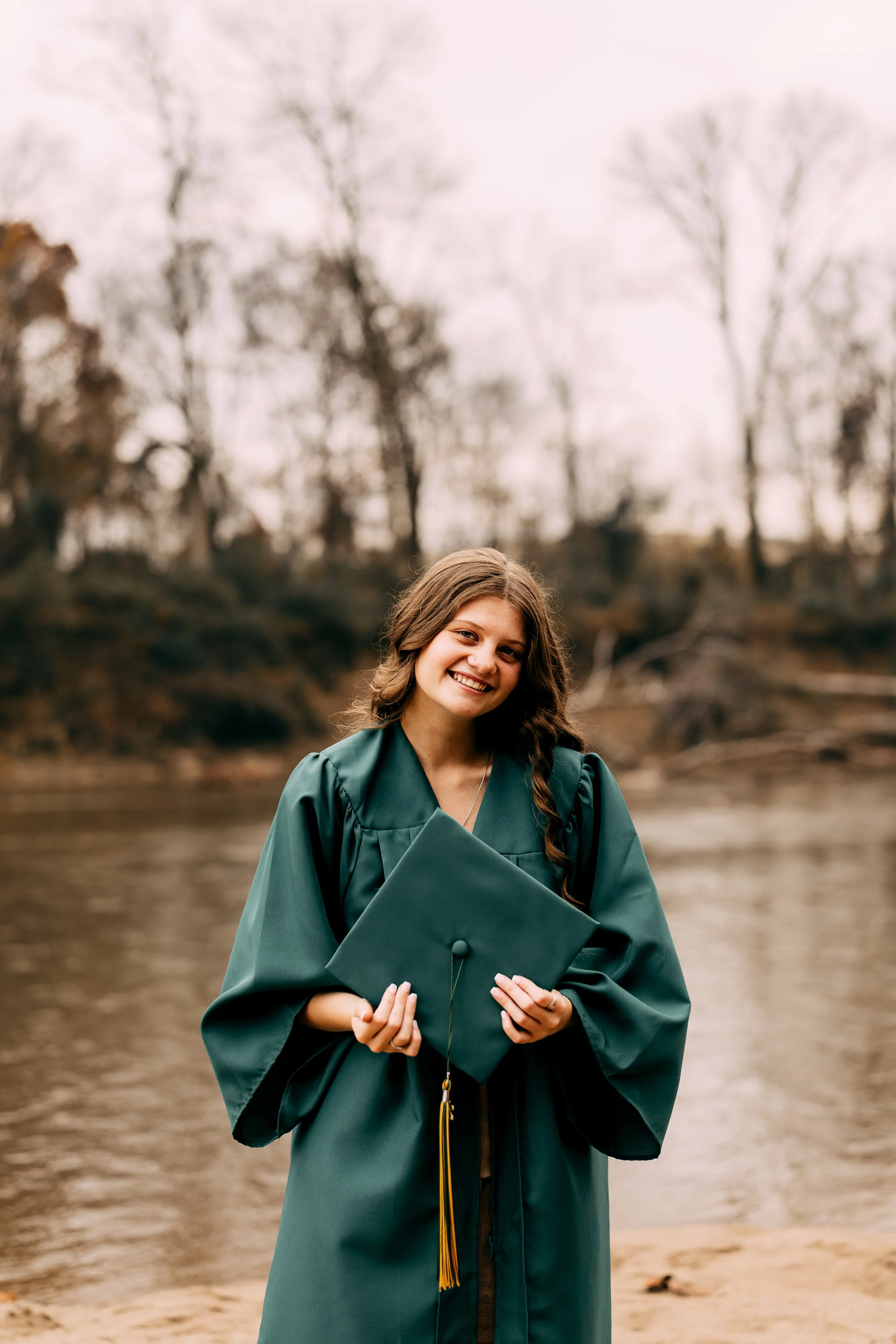A young woman in a green graduation gown holding a cap, smiling outdoors by a river with trees in the background.