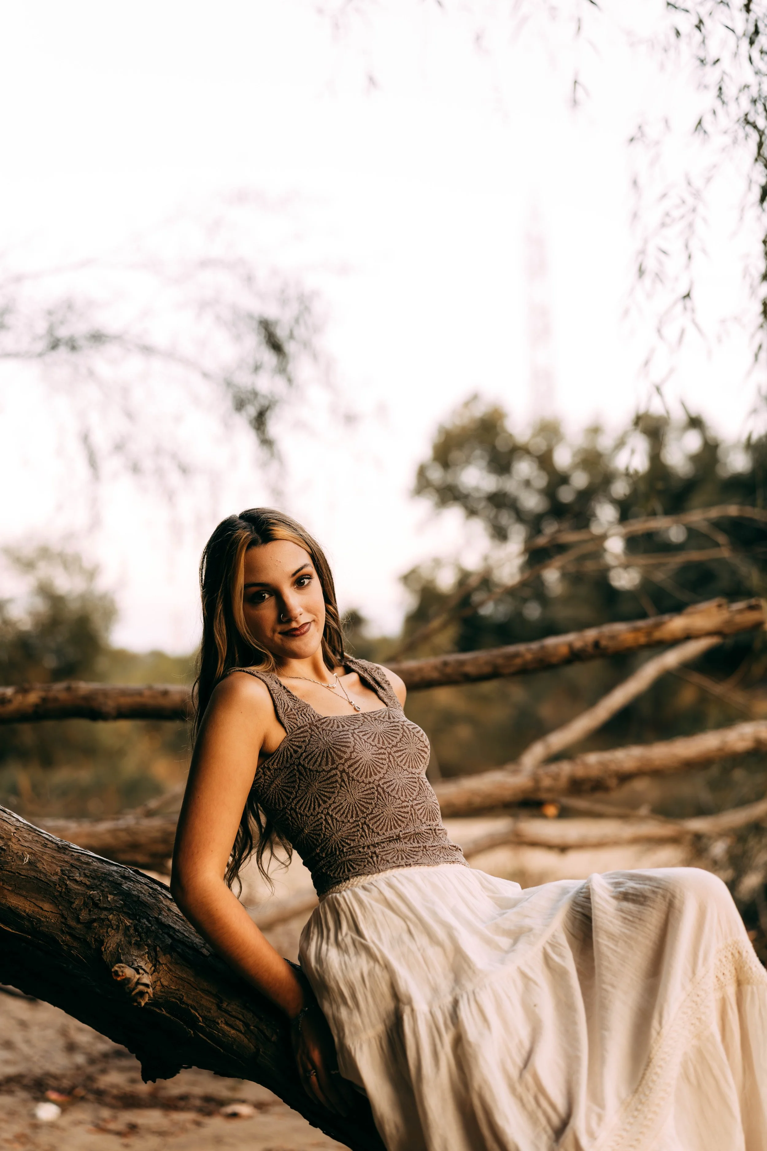 Young woman with long wavy hair sitting outdoors on a fallen tree trunk in a natural setting with trees and branches in the background.