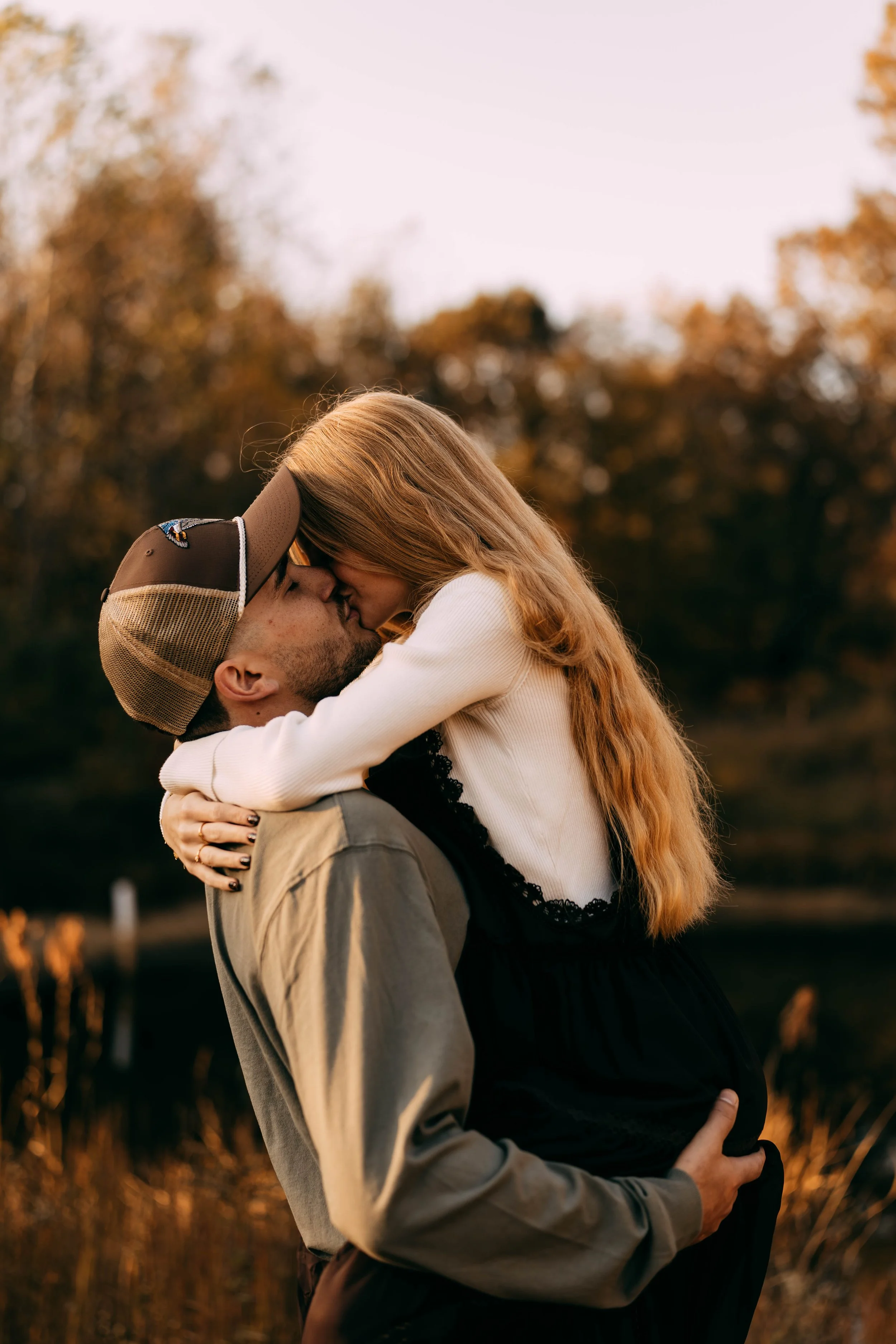 A man wearing a baseball cap and a woman with long red hair share a kiss outdoors during sunset, with autumn trees in the background.