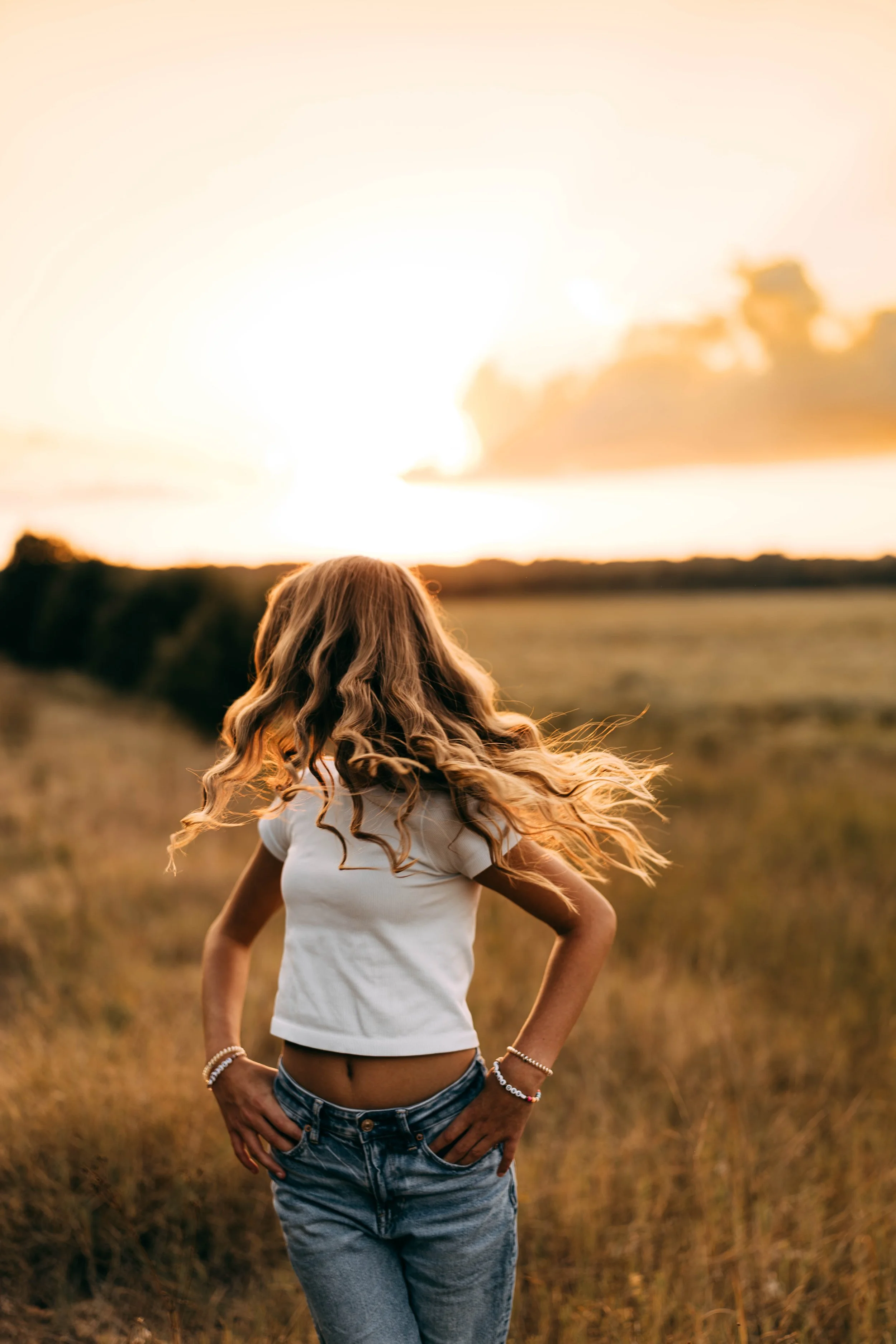 A woman with long, curly hair standing in a field during sunset, facing away from the camera.
