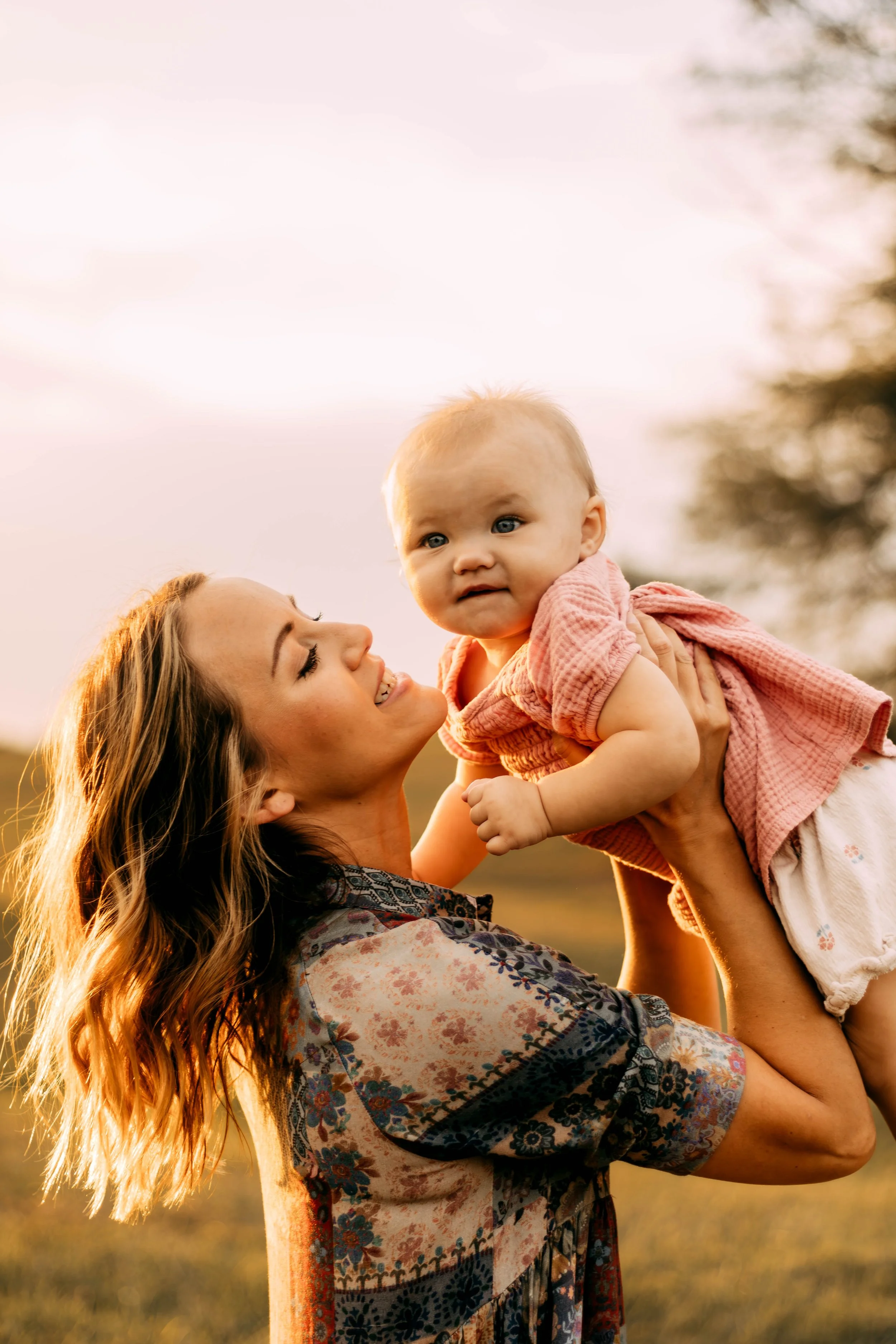 A woman with brown wavy hair holding a baby girl outside during sunset. The woman is smiling and the baby is looking at the camera with a curious expression.
