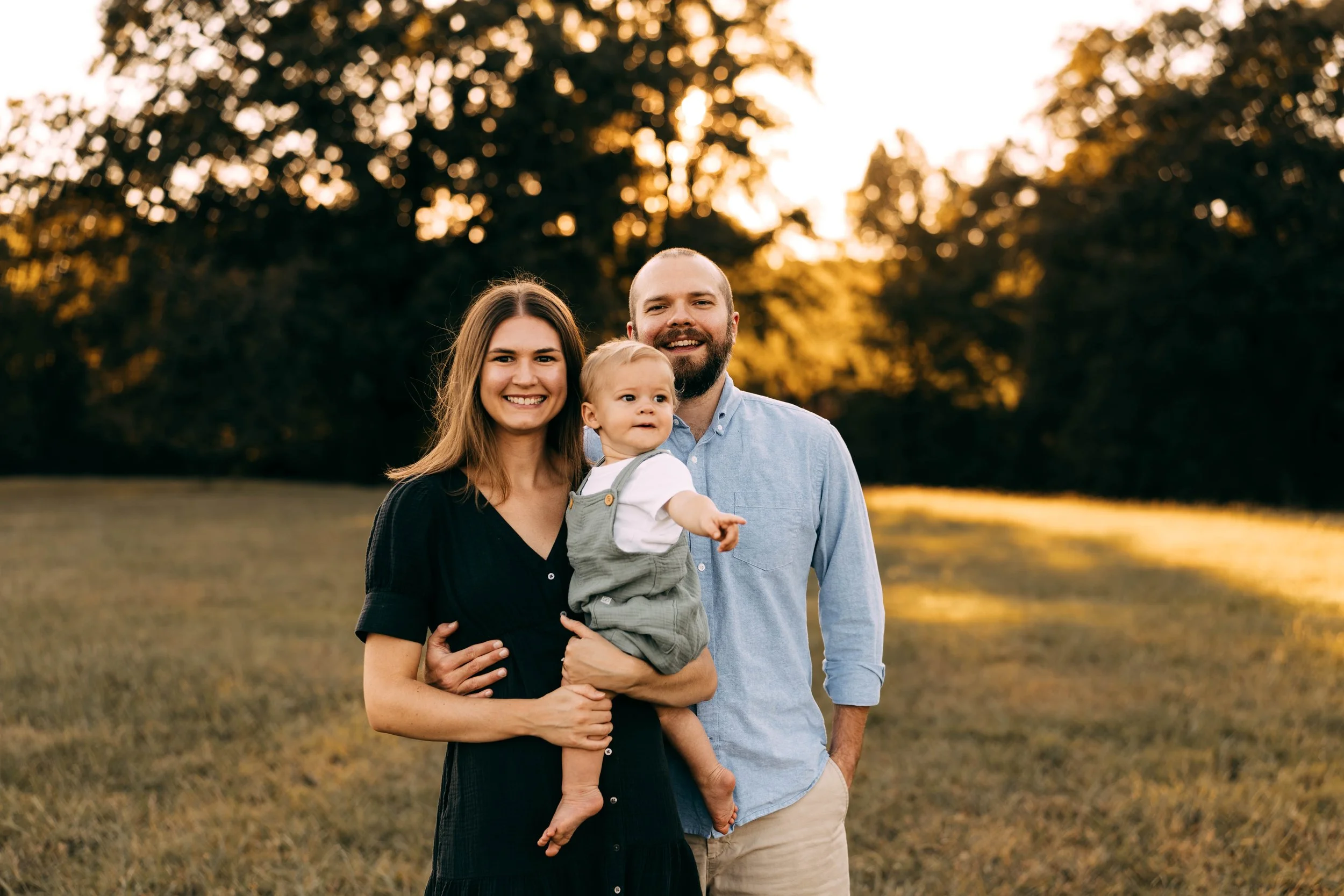 A happy family consisting of a woman, a man, and a young child, standing outdoors in a field during sunset, smiling at the camera.
