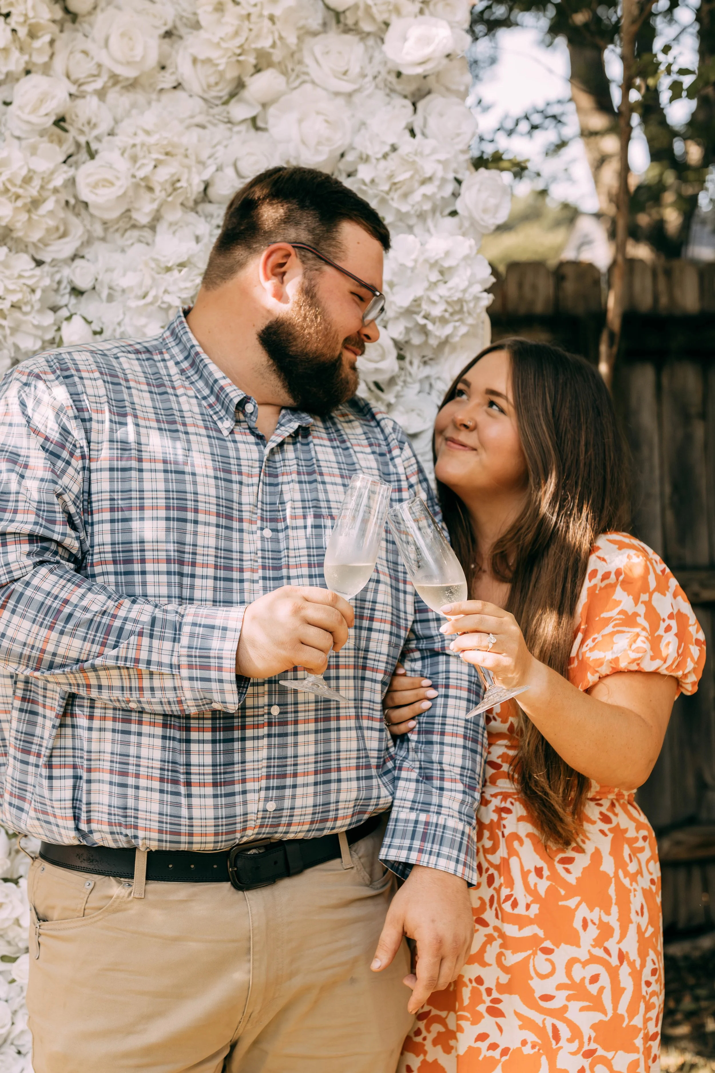 A couple celebrating outdoors, holding champagne glasses, standing in front of a white floral backdrop.