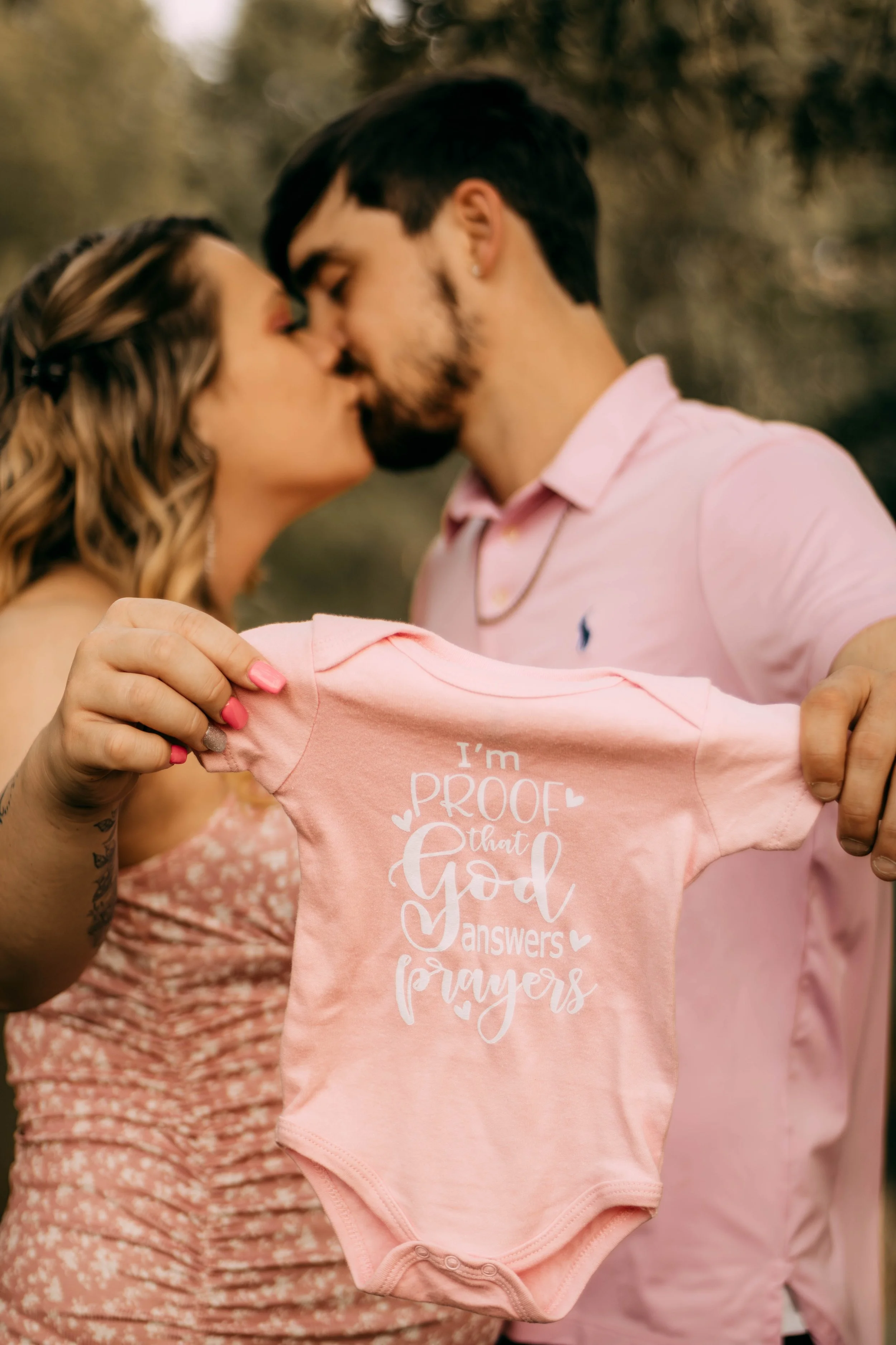 A couple kissing outdoors while holding up a pink baby onesie that says, 'I'm proof that God answers prayers.'