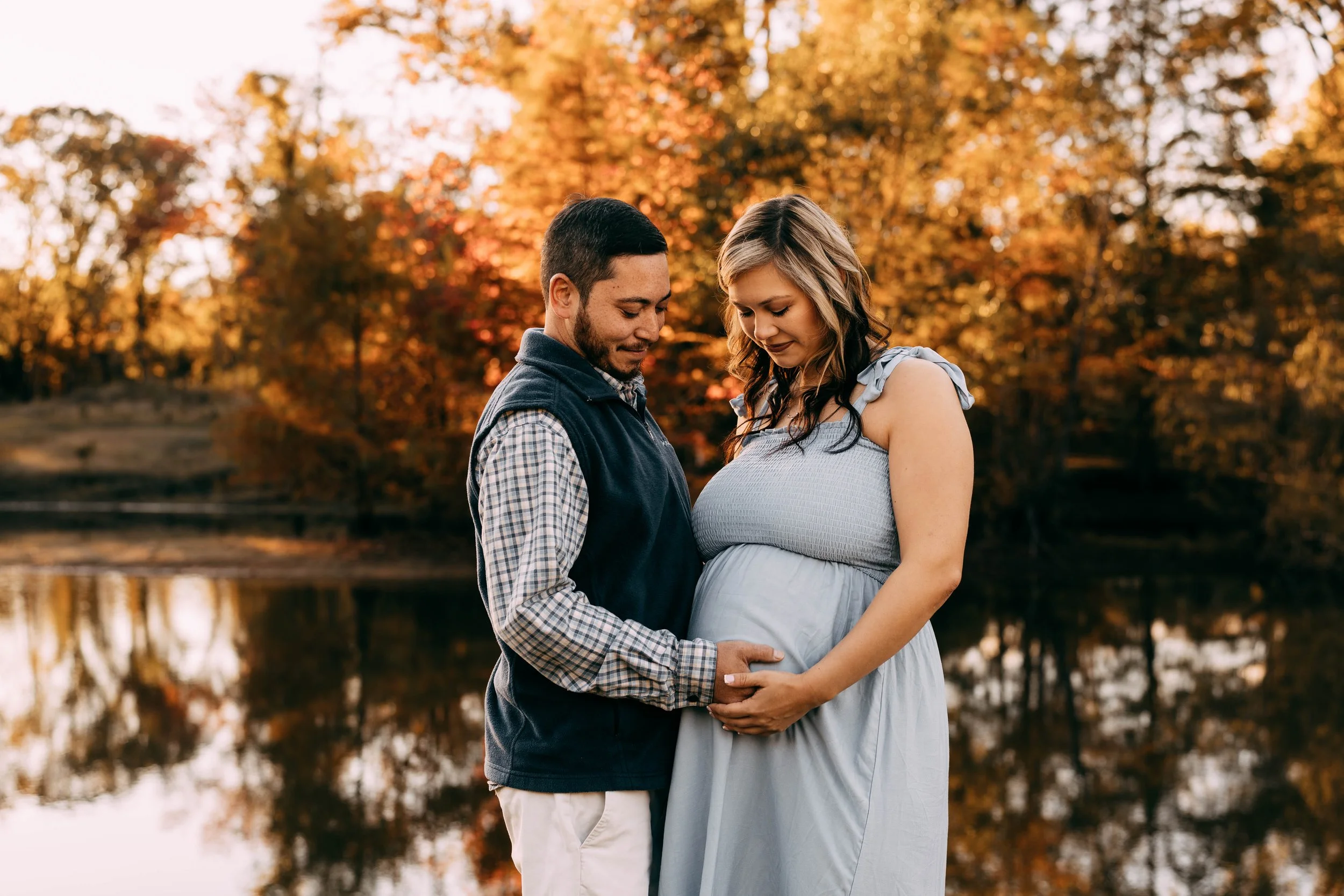 Pregnant woman and man standing by a lake during autumn, holding her belly and looking down, surrounded by fall foliage.