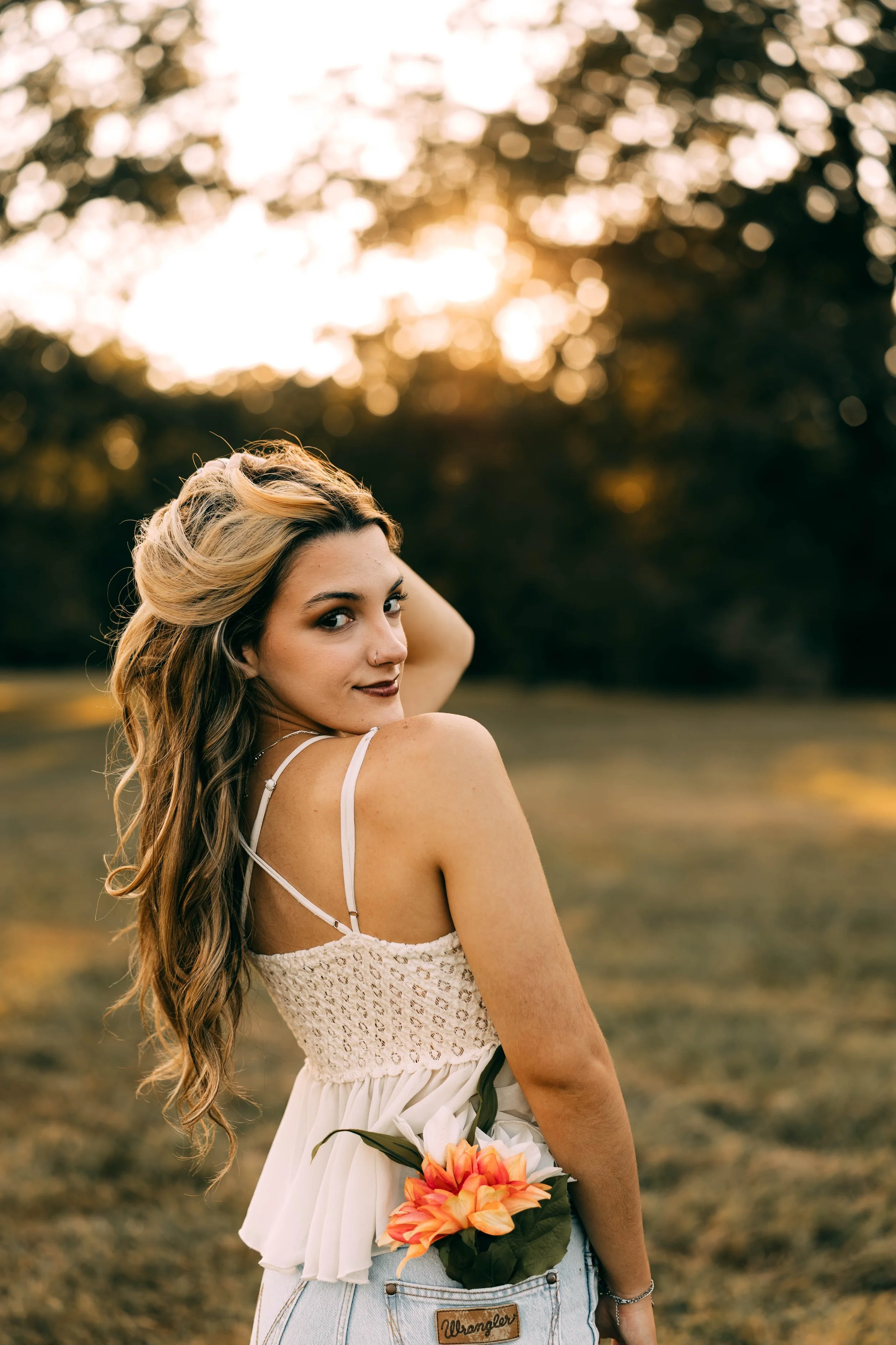 A young woman with long, wavy hair standing outdoors during sunset, wearing a white top and holding a pink and orange flower in her back pocket.
