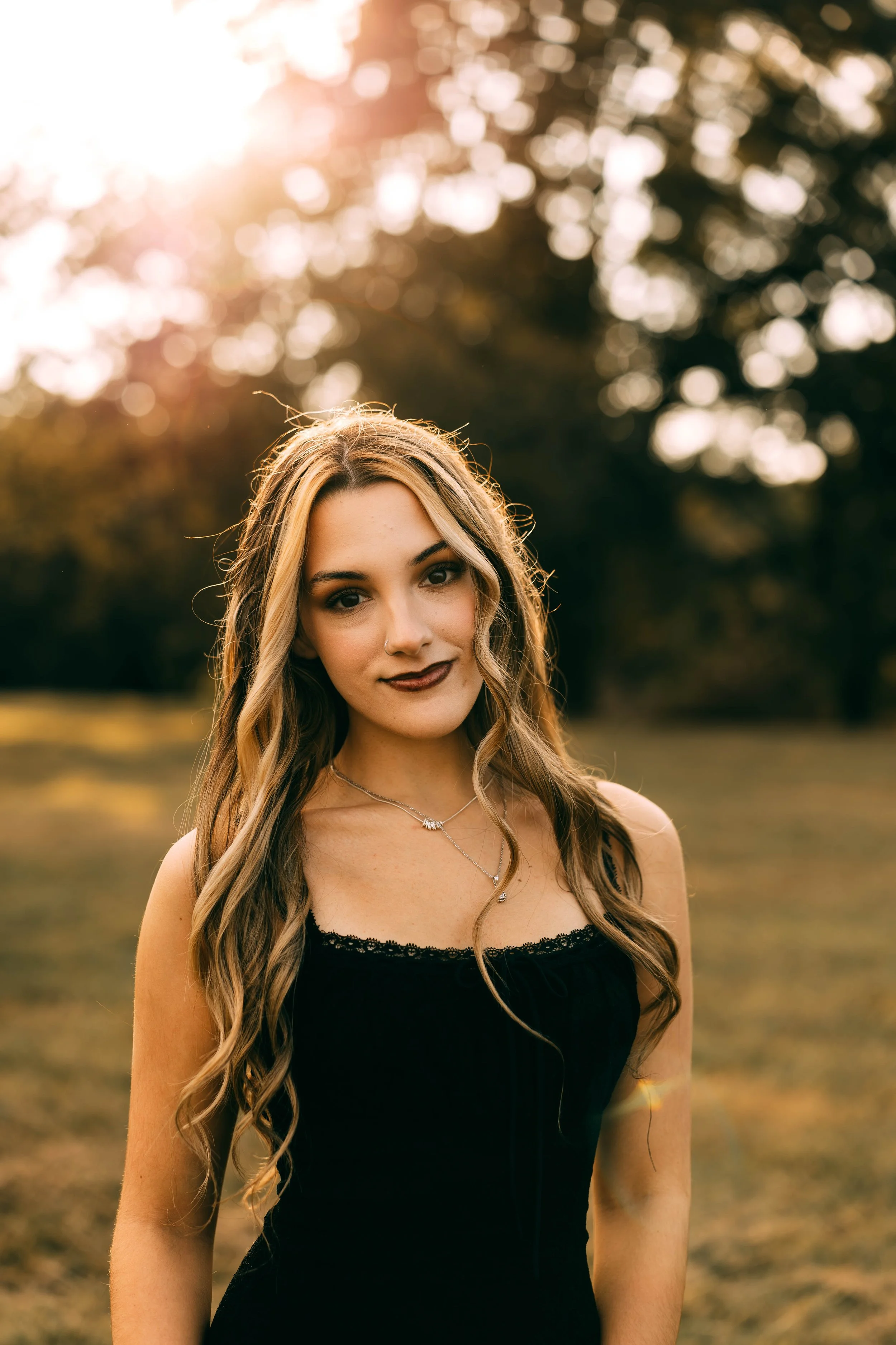 A young woman with long wavy hair in a black top outdoors during sunset, with sun flare and blurred trees in the background.