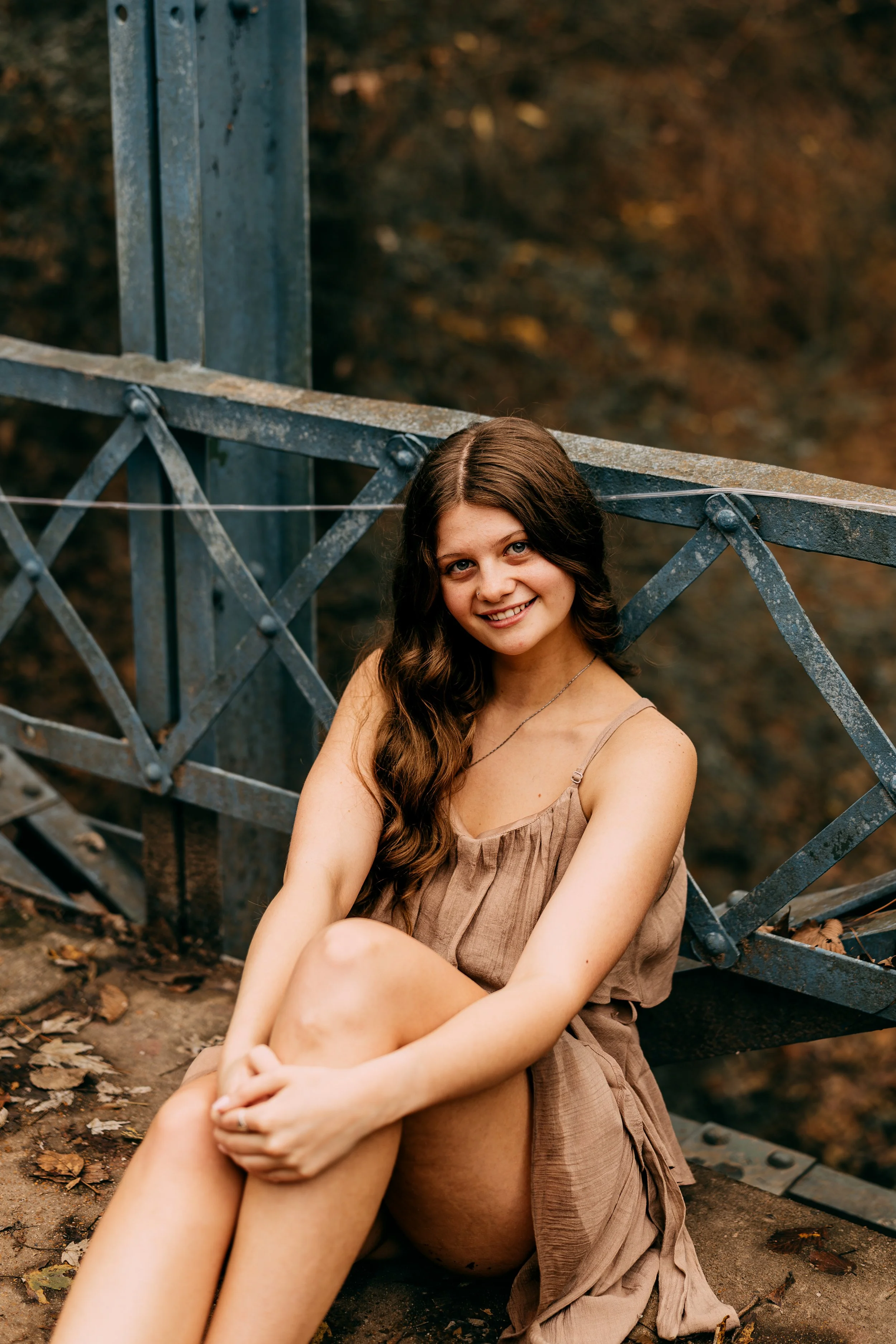 Young woman with long, wavy brown hair sitting on the ground near a metal railing, smiling at the camera with a blurred autumn forest background.