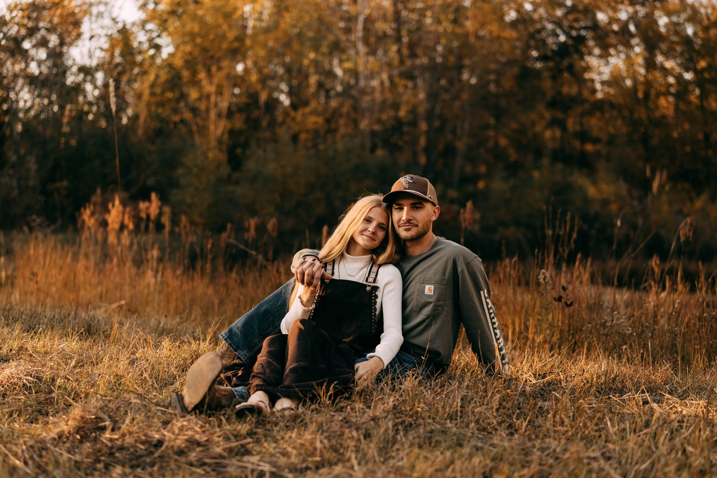 Young couple sitting on grass in a field during autumn sunset, outdoors, surrounded by trees with fall foliage.