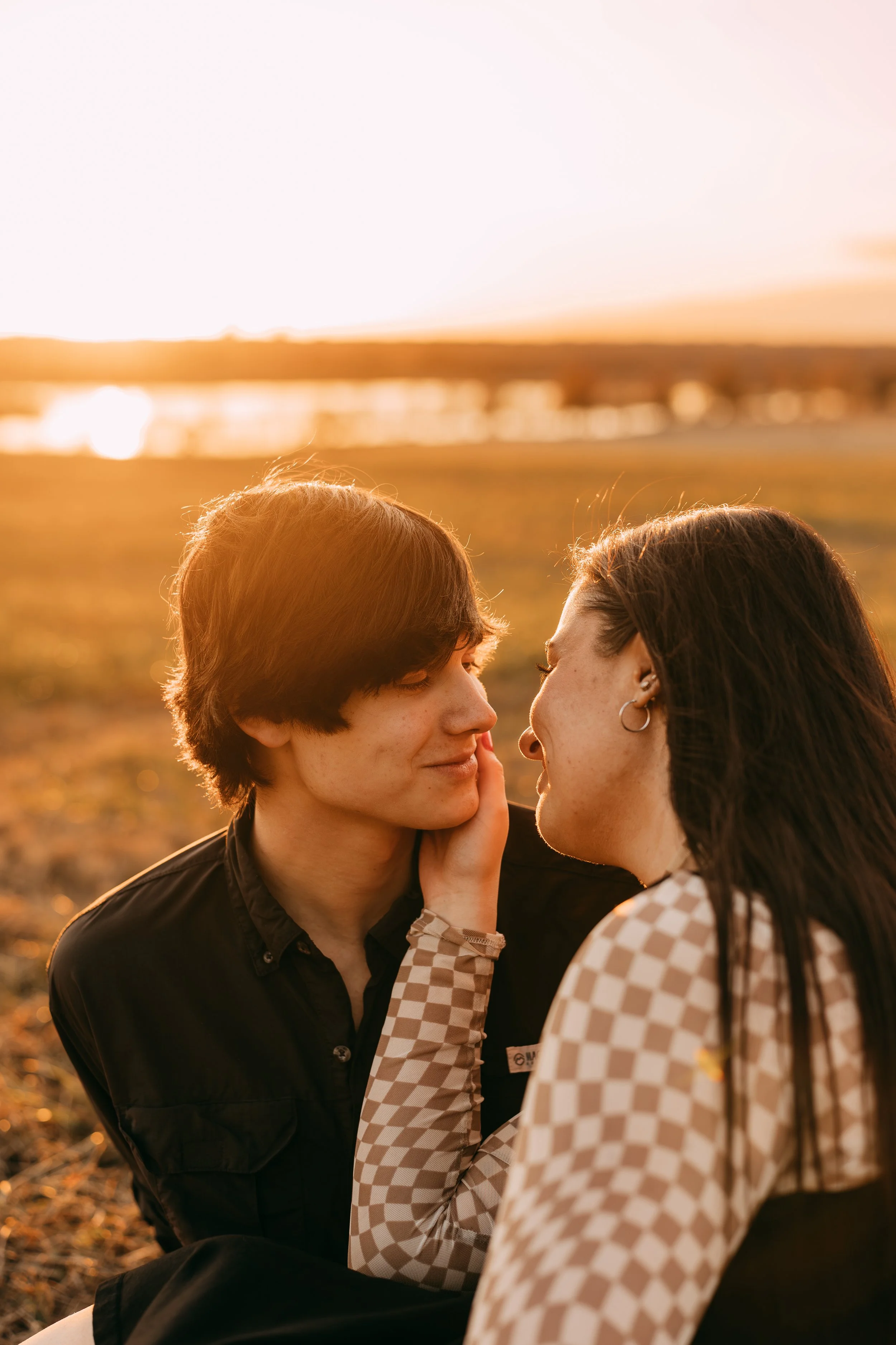 A couple sitting close together outdoors at sunset, with water and a sunset sky in the background. The woman is gently touching the man's cheek as they gaze at each other.