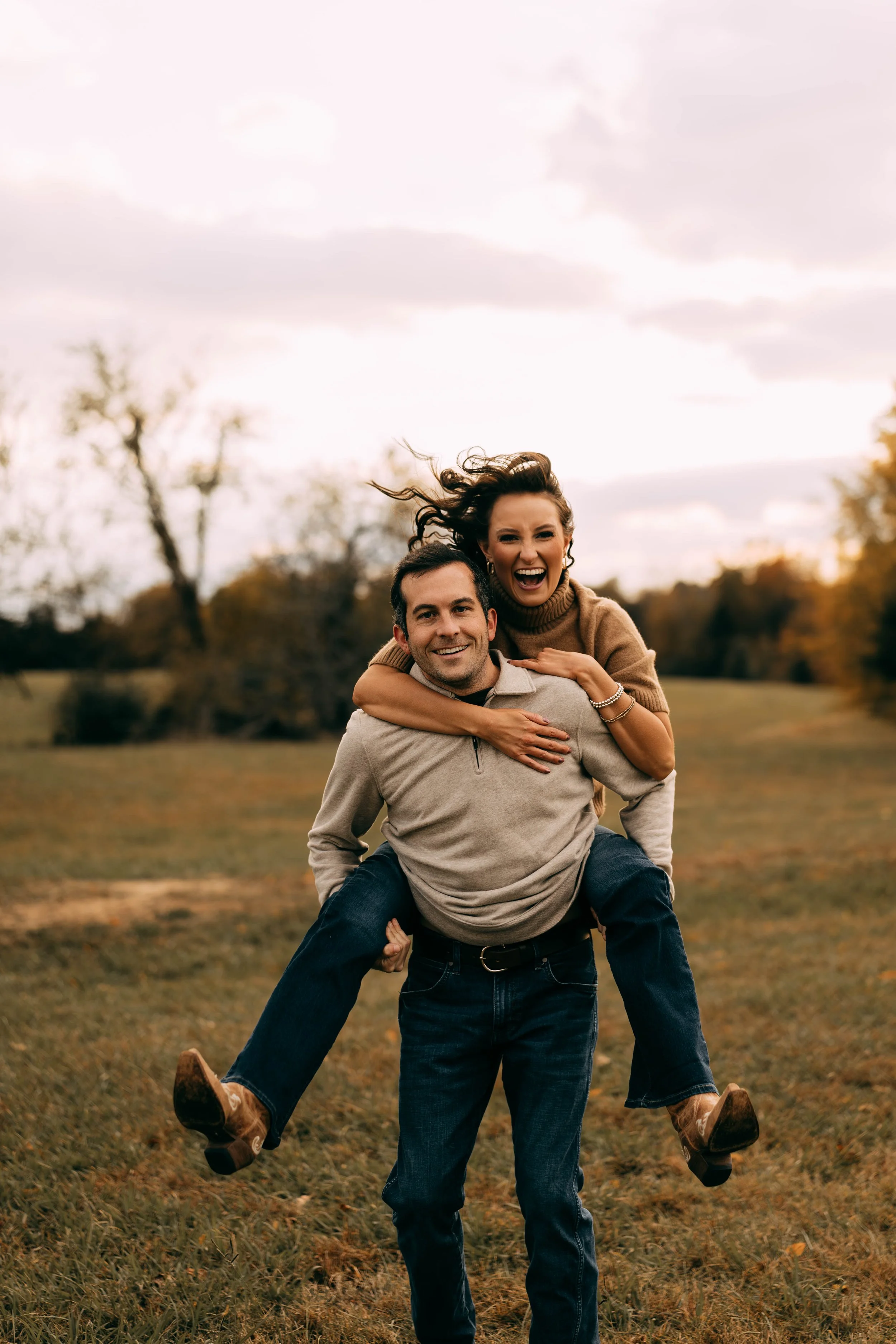 A happy couple in a park during fall, with the woman on the man's back, both smiling and enjoying the moment.