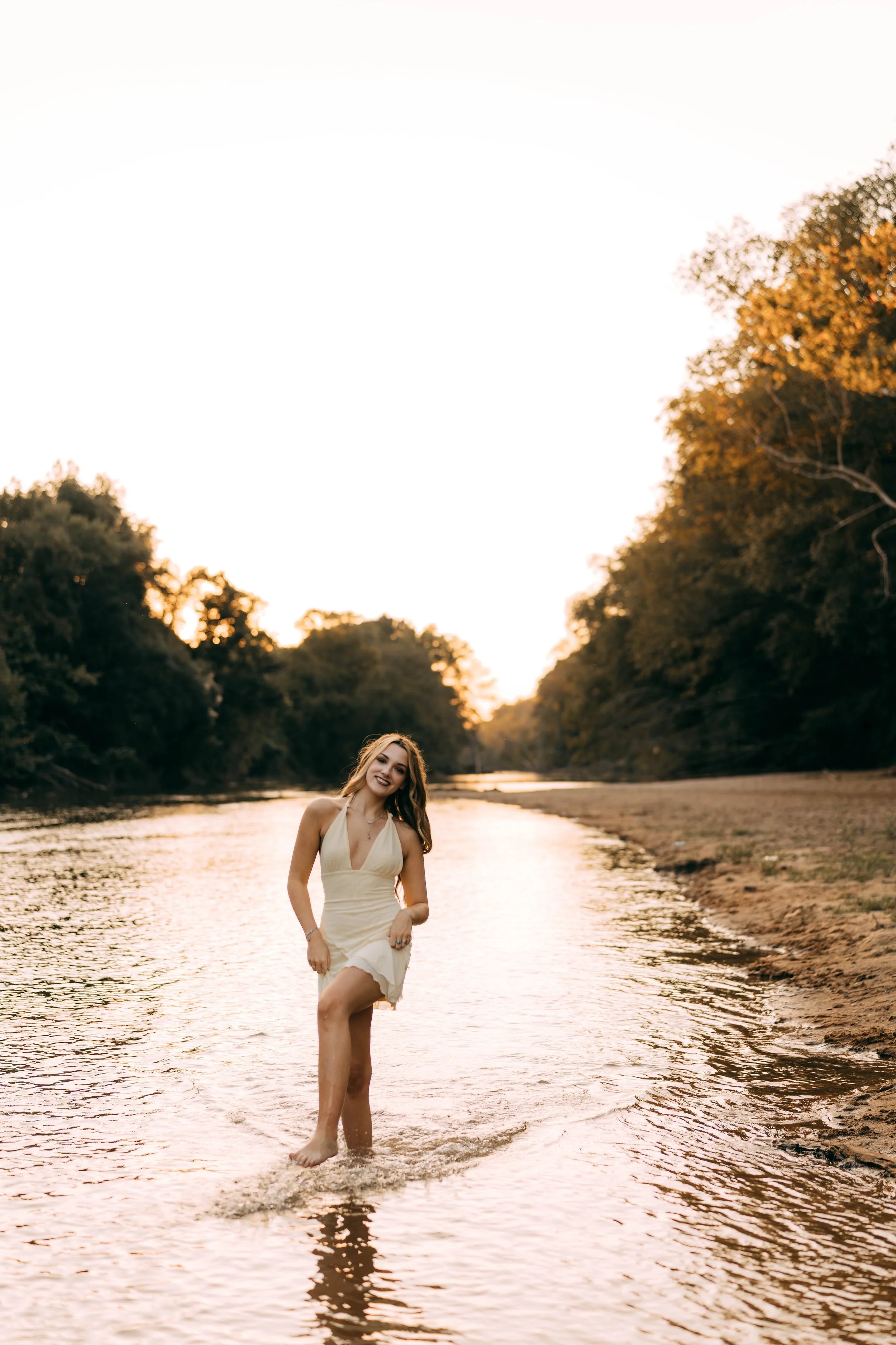 A young woman in a beige dress standing in a shallow river at sunset, smiling and looking at the camera, with trees on both sides of the river.