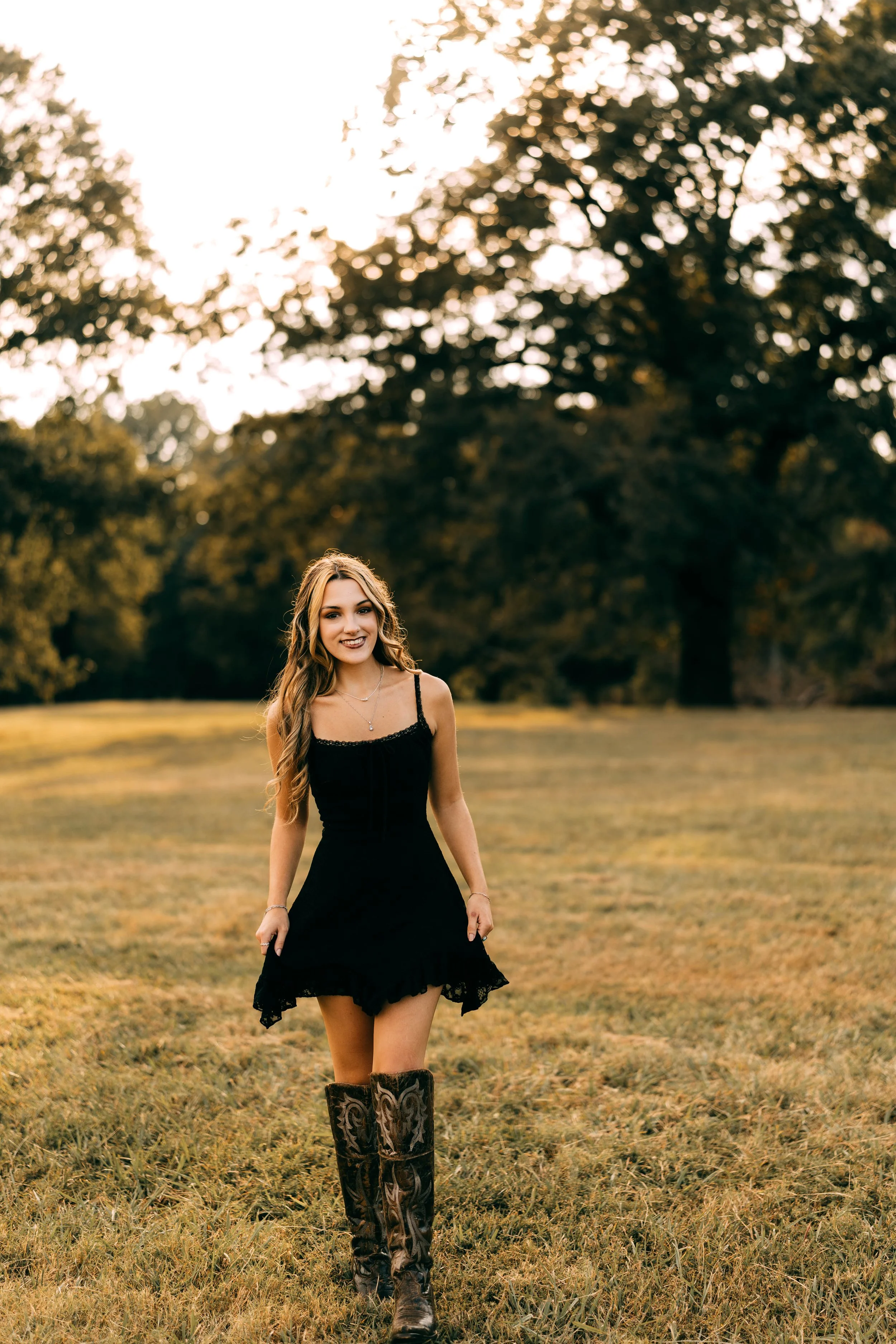 A young woman with long wavy hair wearing a black dress and cowboy boots walking on a grassy field with trees in the background during sunset.