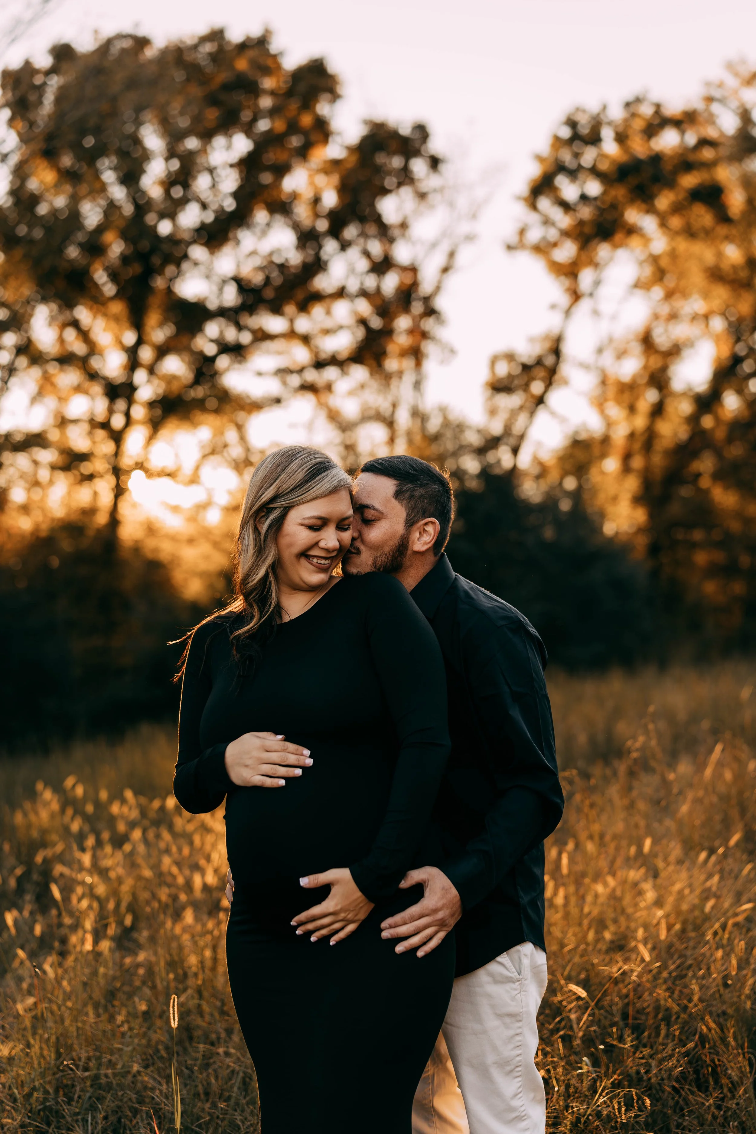 A pregnant woman and a man are in a field during sunset, with trees in the background. The woman is smiling and wearing a black long-sleeve dress, while the man is kissing her cheek and wearing a black shirt and light-colored pants.