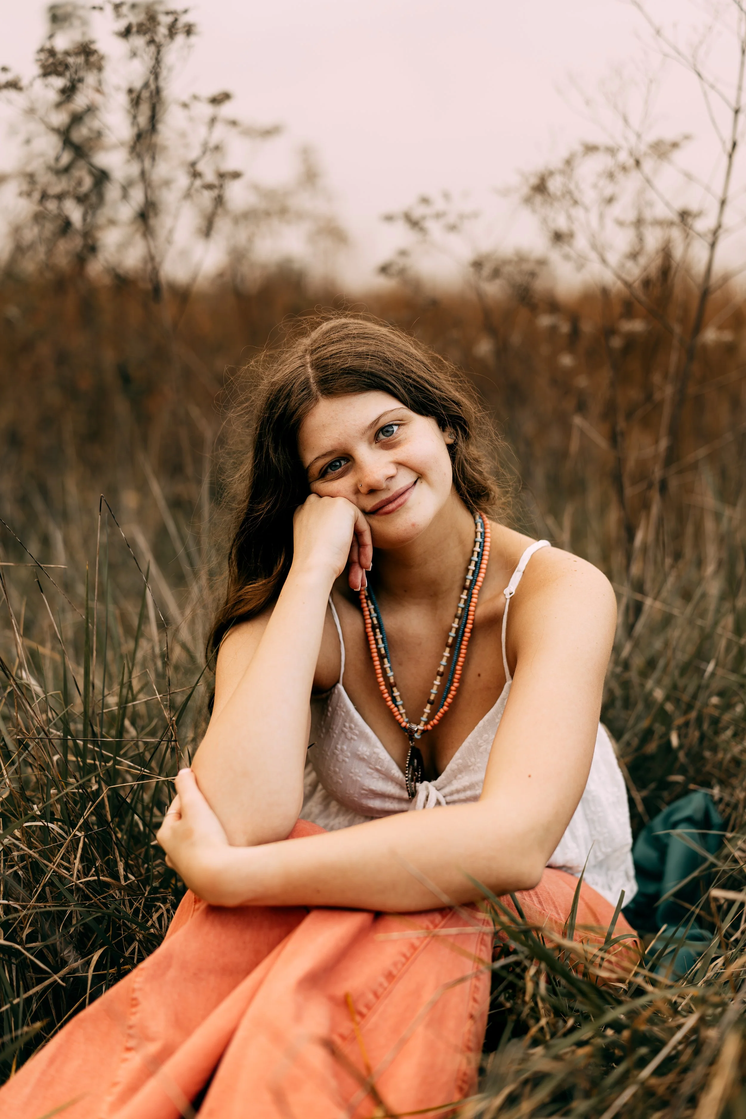 A young woman sitting in a field of dry grass, smiling at the camera with her head resting on her hand. She has long brown hair, blue eyes, and is wearing a white top, orange skirt, and beaded necklaces.