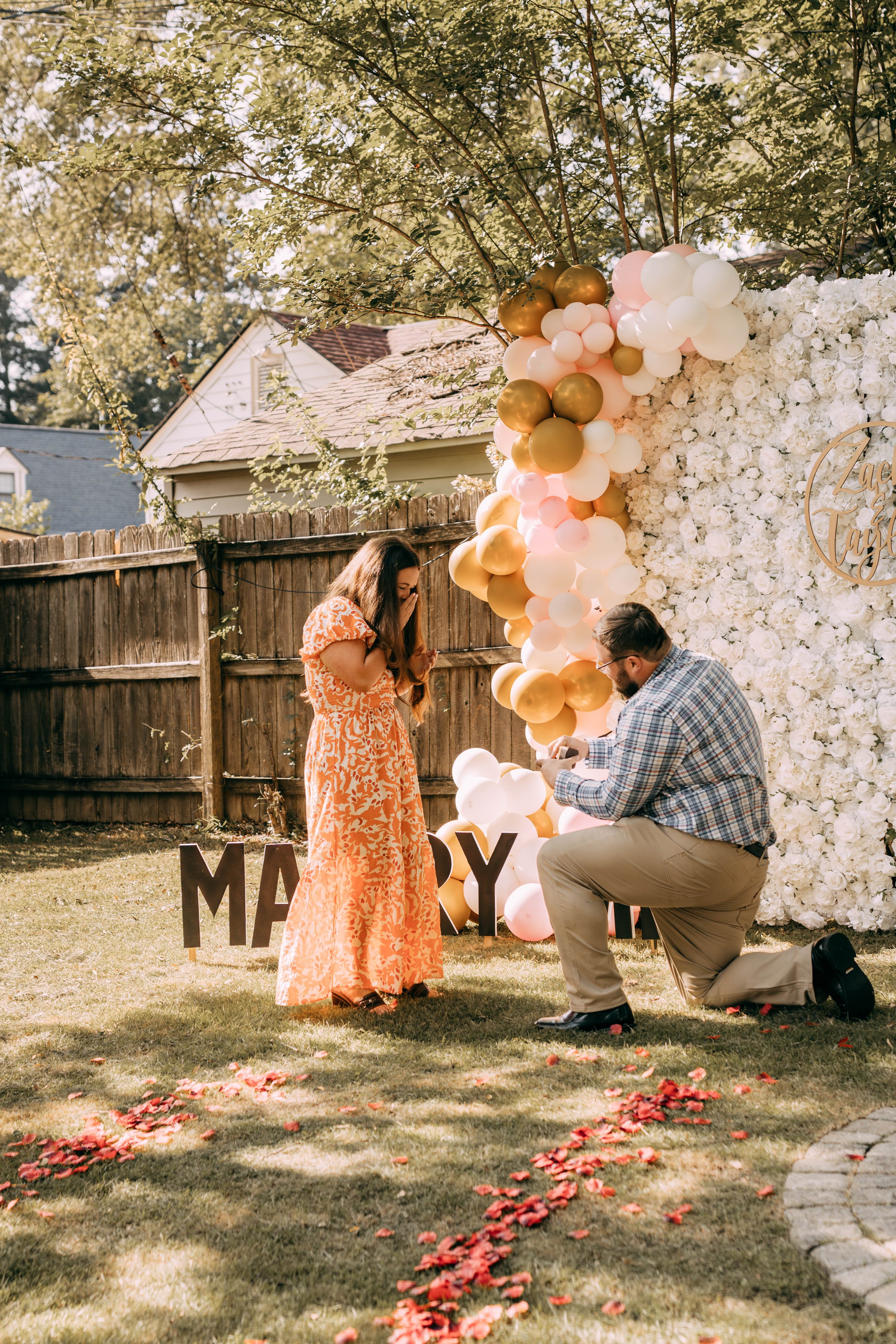 A man proposing to a woman in a backyard wedding or engagement setting with floral and balloon decorations.