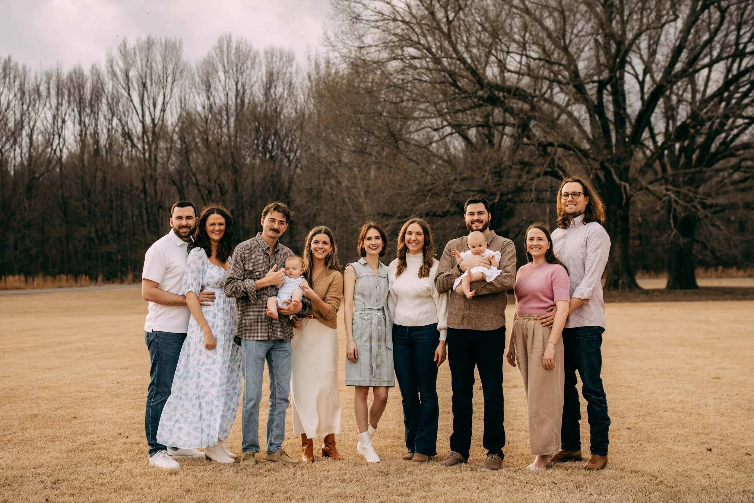 A group of eleven people, including two babies, standing outdoors on a grassy field with leafless trees in the background, smiling at the camera.