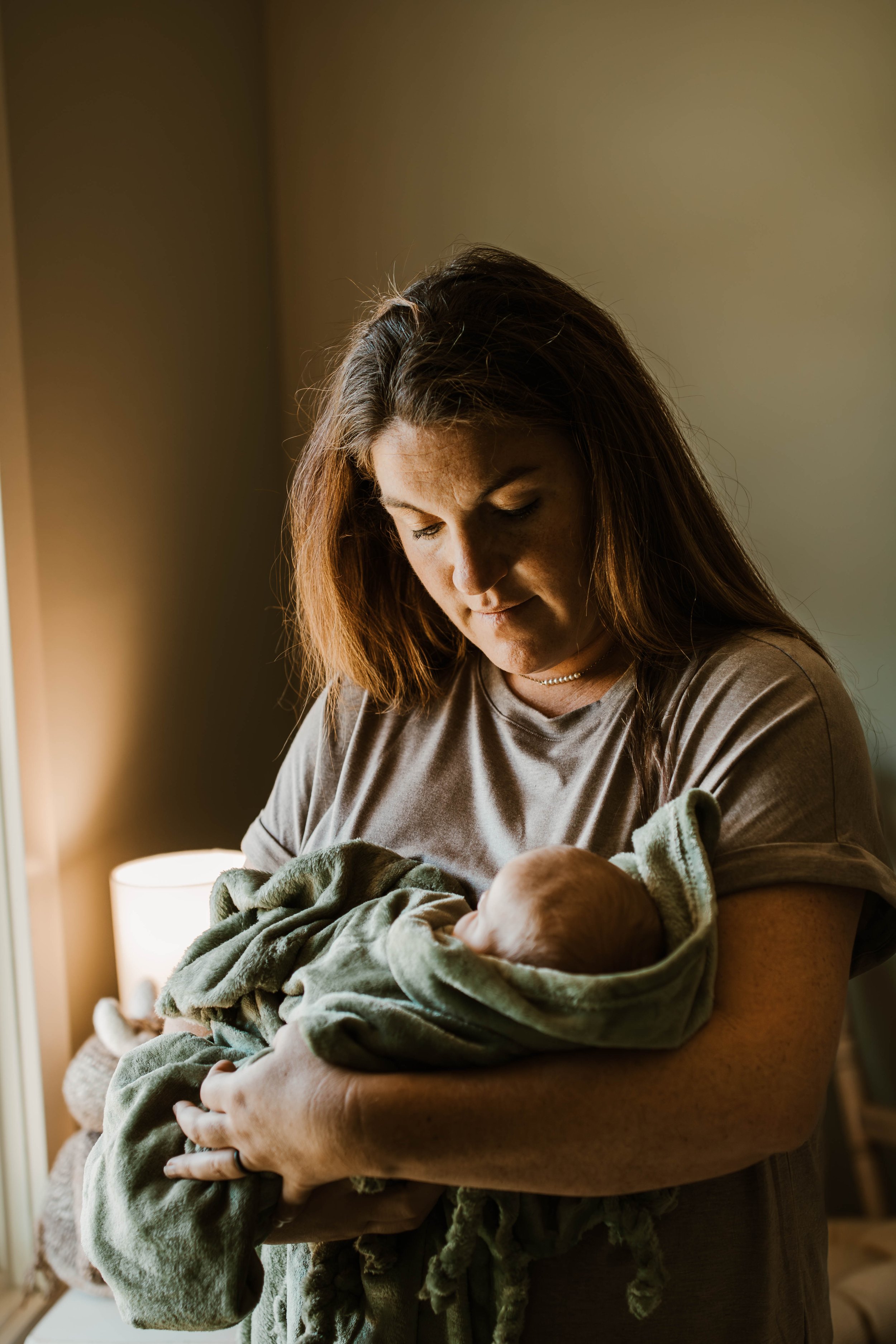 A woman with brown hair holding a newborn wrapped in a green blanket in a softly lit room.