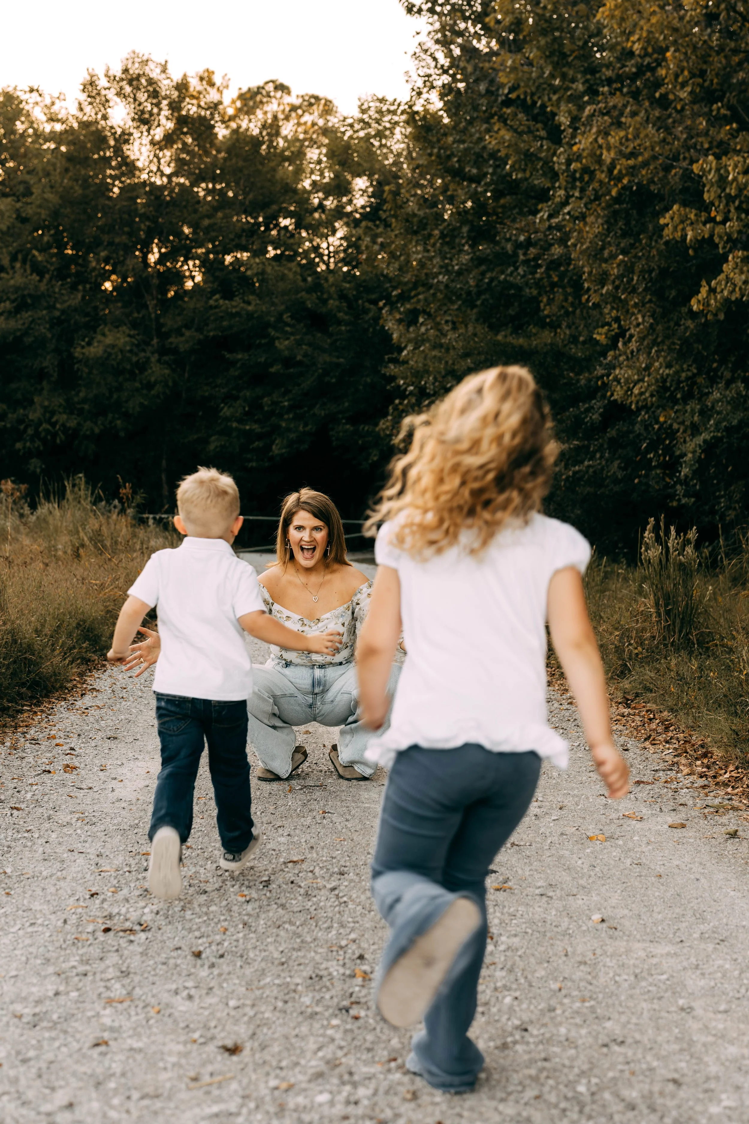 A woman with shoulder-length hair in a floral top crouching and smiling as two children run towards her on a gravel path in a park at sunset, surrounded by trees with green foliage.