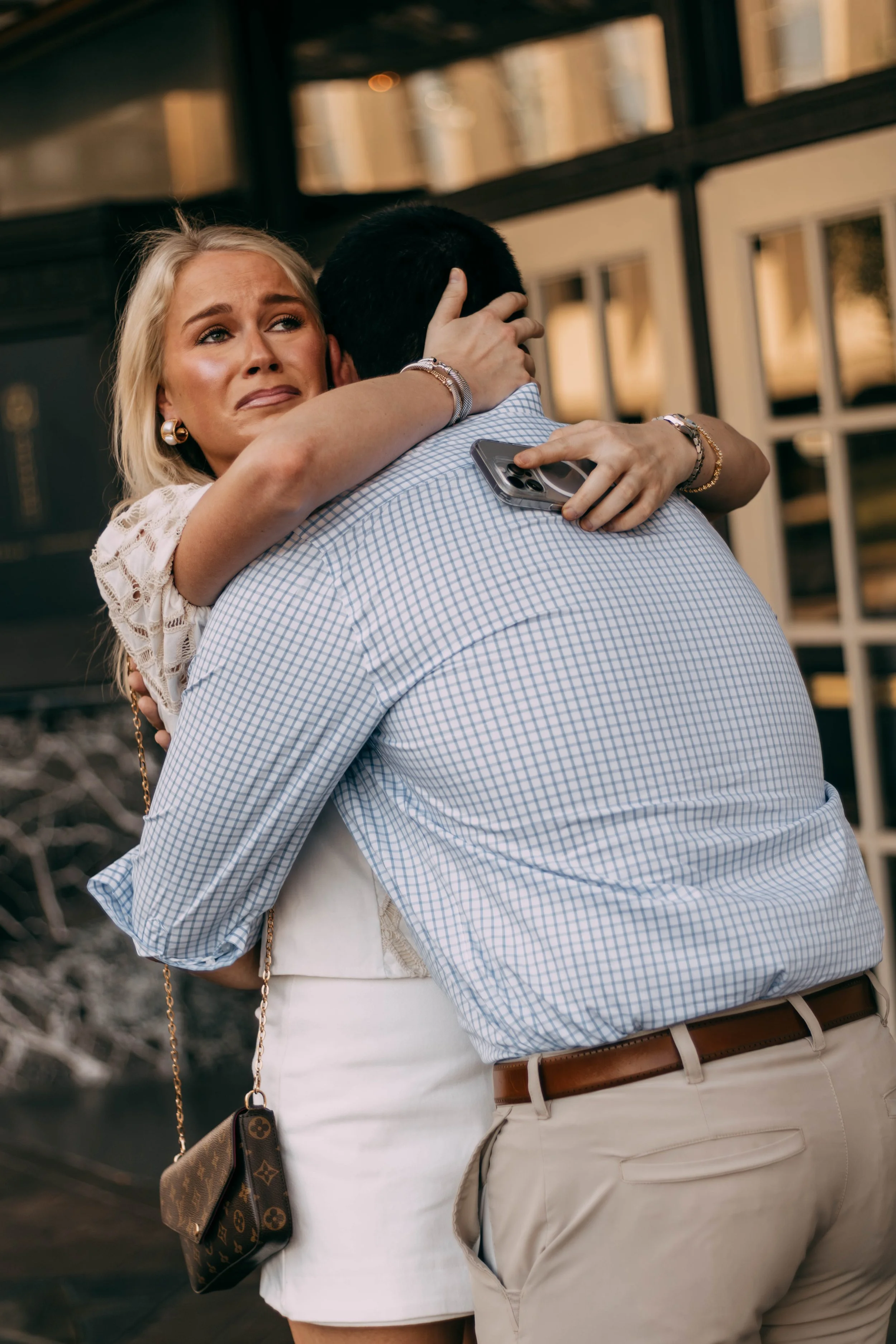A woman with blonde hair embraces a man, visibly distressed, outdoors near a building with glass windows. The woman holds a smartphone in her hand.