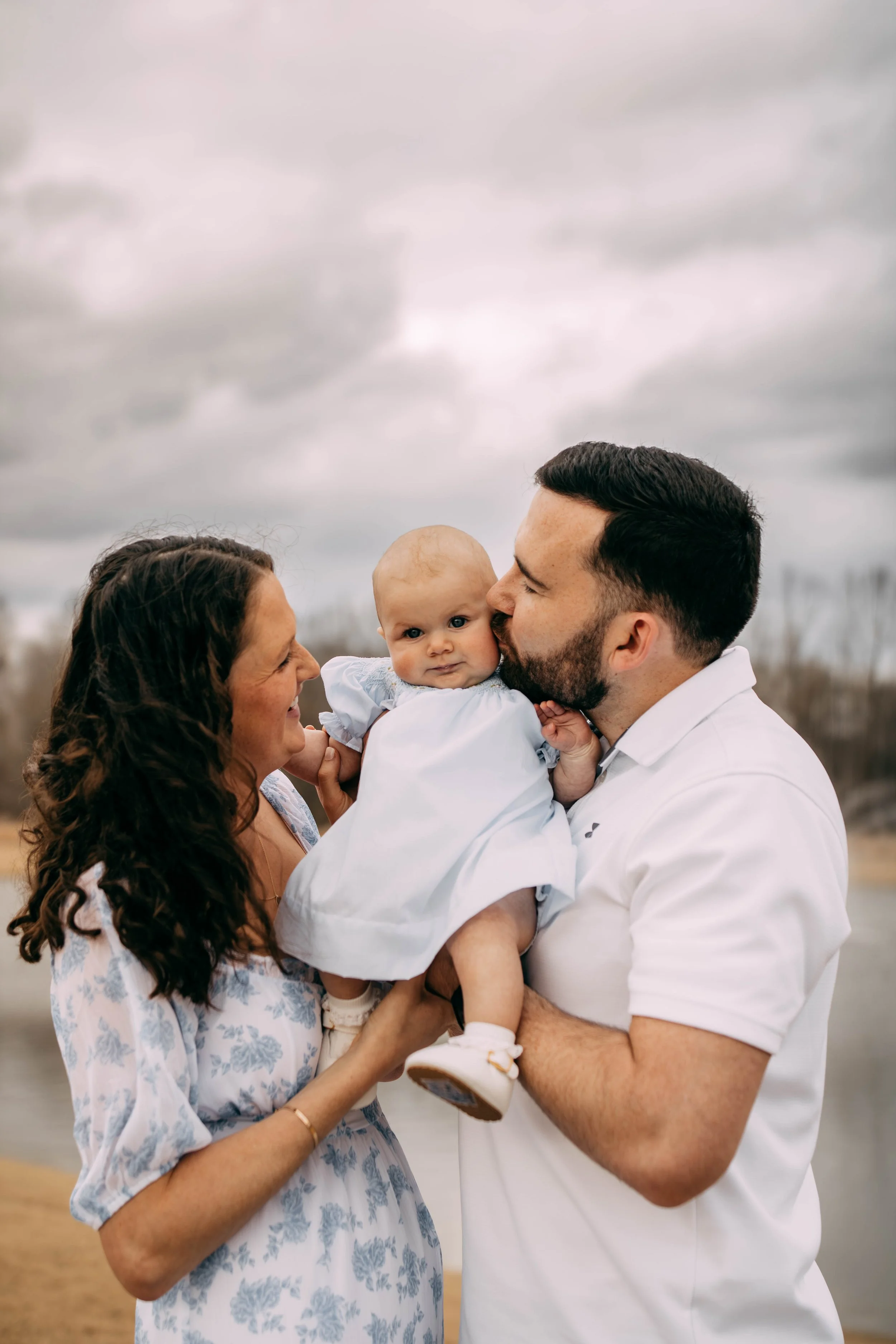 Family of three outdoors, mother, father, and a baby girl, with a cloudy sky in the background. The mother has curly hair and is smiling at the father, who is kissing the baby on the cheek. The baby girl has a serious expression, is dressed in a whit