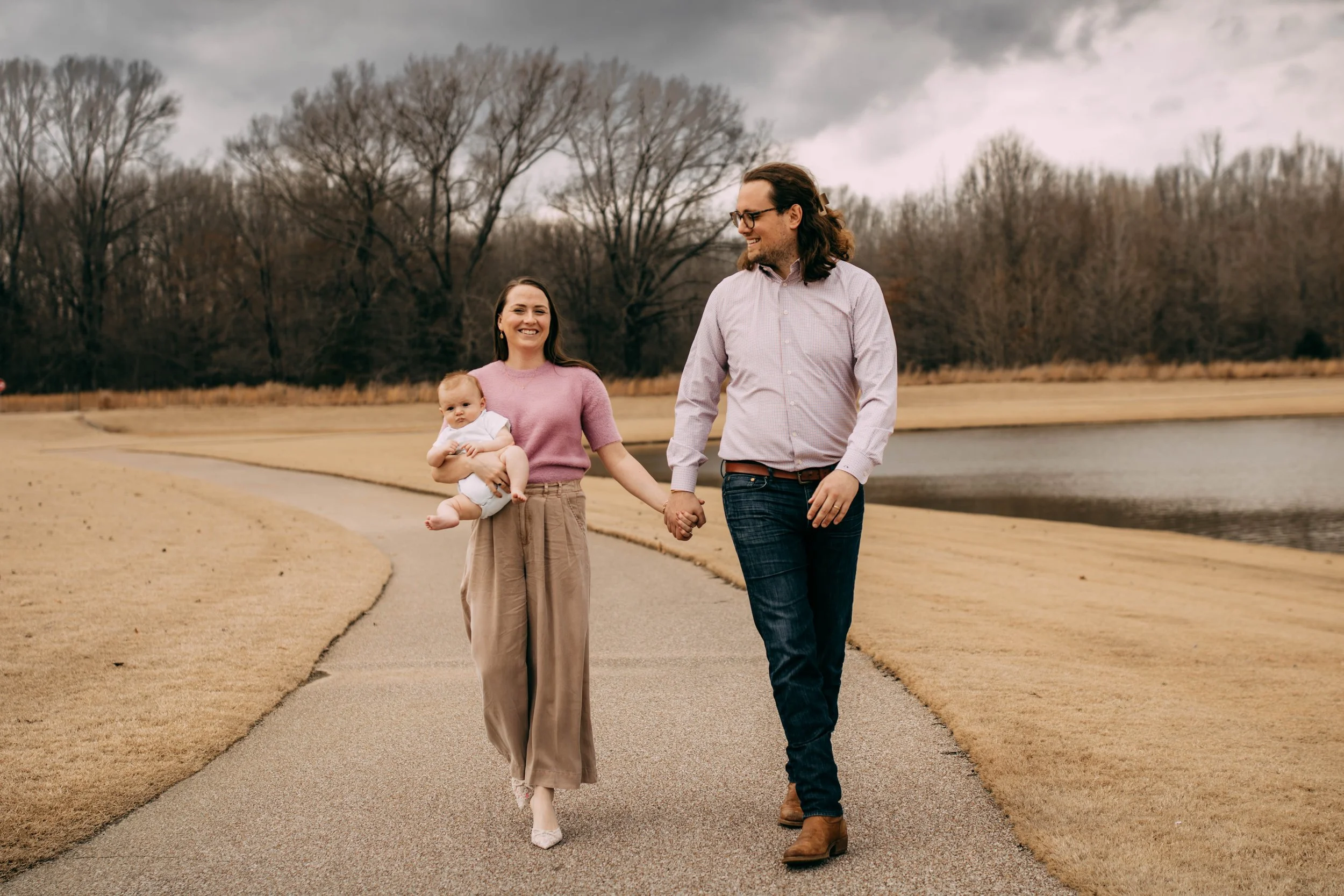 A family of three walking on a paved path by a pond, holding hands and smiling, with leafless trees in the background.
