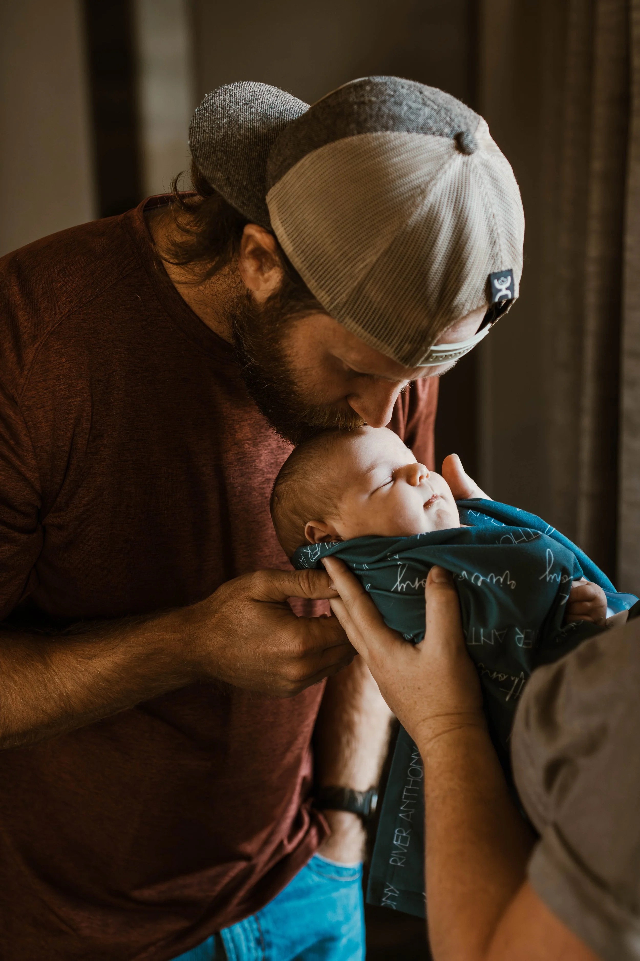 A man with a beard and cap holding a sleeping newborn baby wrapped in a blue blanket.
