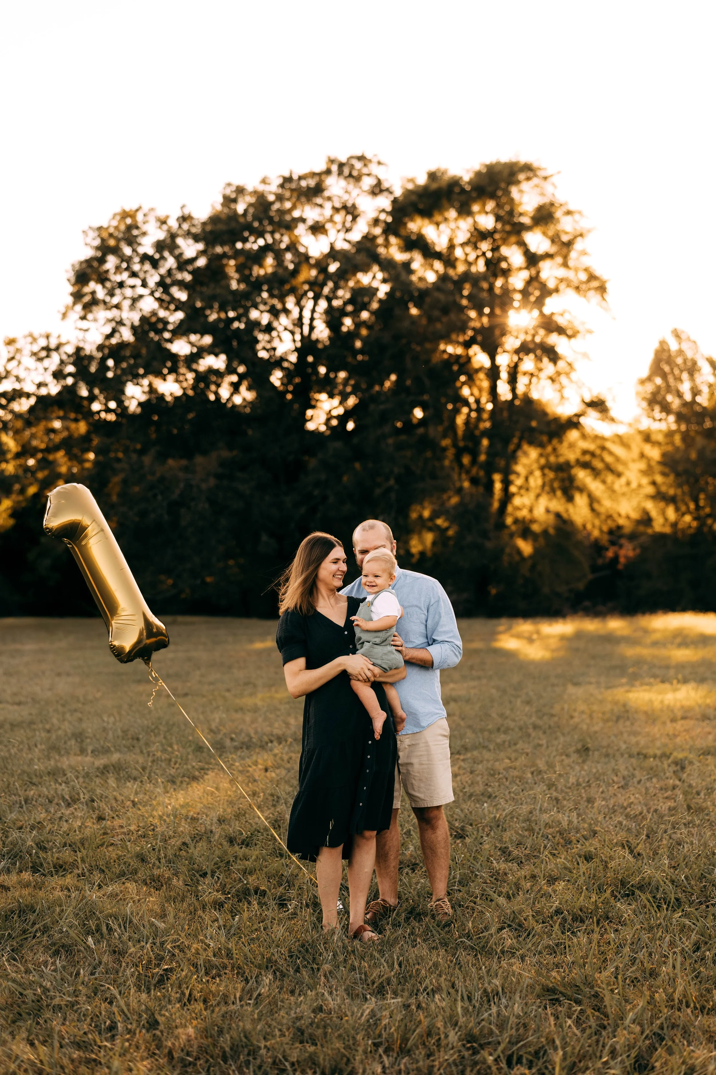 A family of three standing in a field at sunset, with a large golden balloon shaped like the number one.