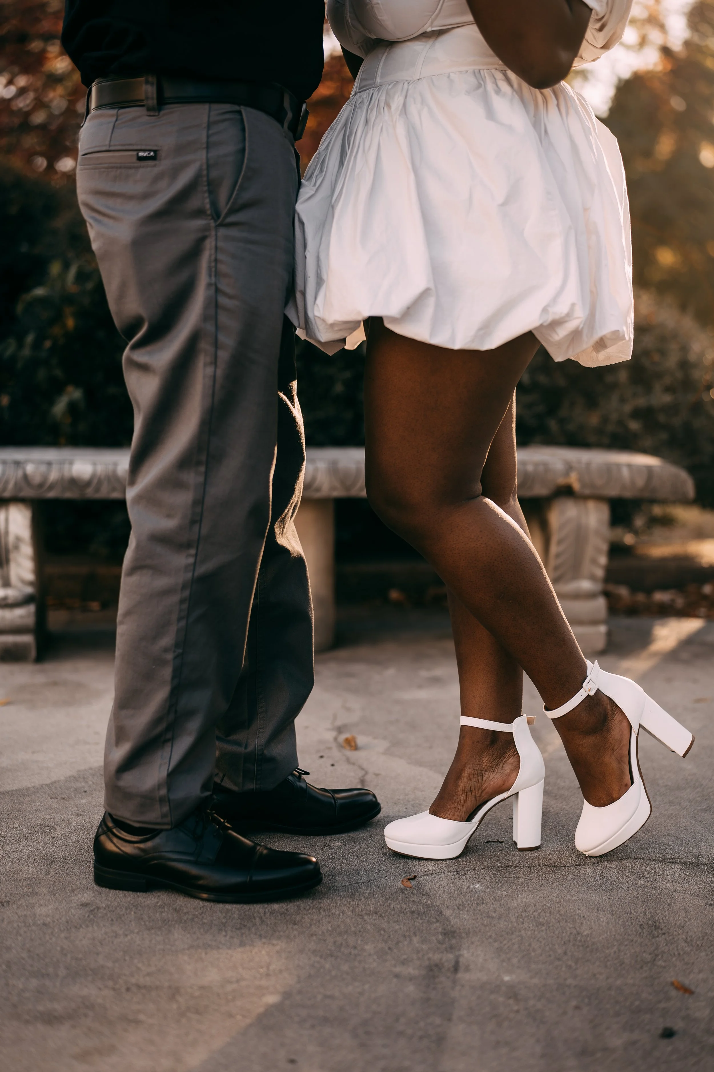 Close-up of two people standing close together outdoors, focusing on their legs and feet. One person is wearing gray dress pants and black shoes, while the other is wearing a white puffed skirt, black tights, and white high-heeled shoes. It appears t