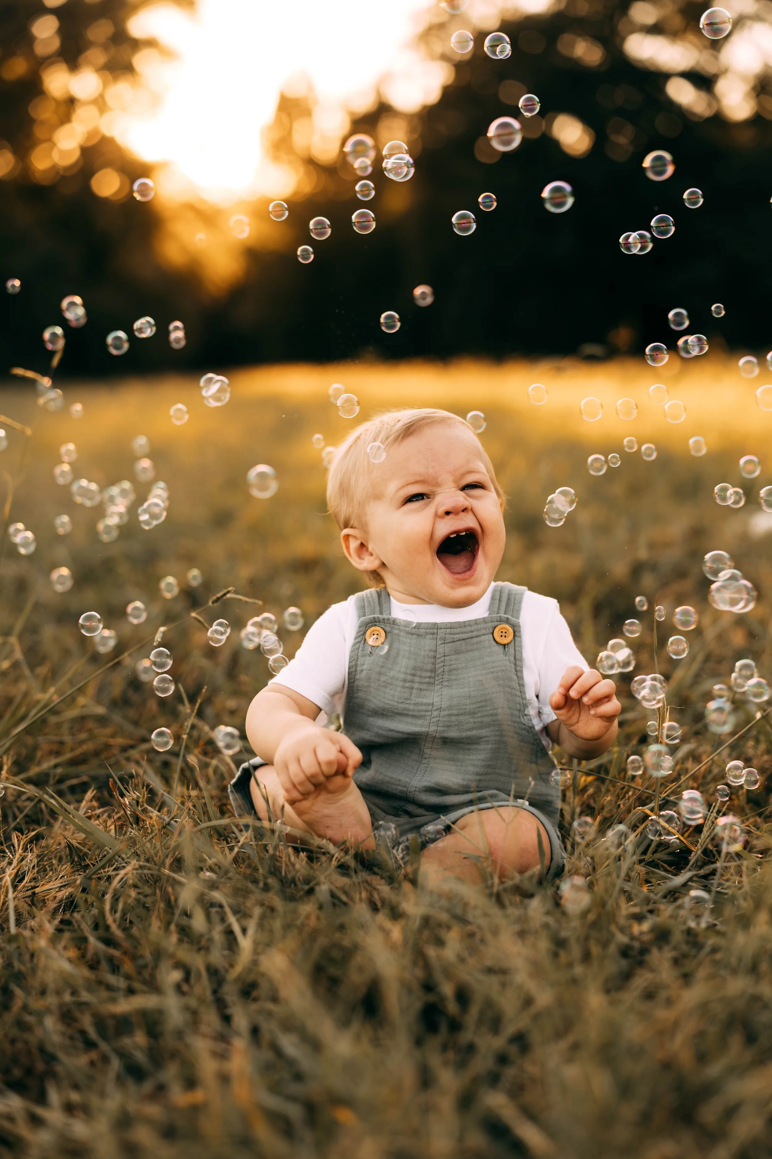 A toddler boy sitting on grass surrounded by bubbles during sunset at a park.