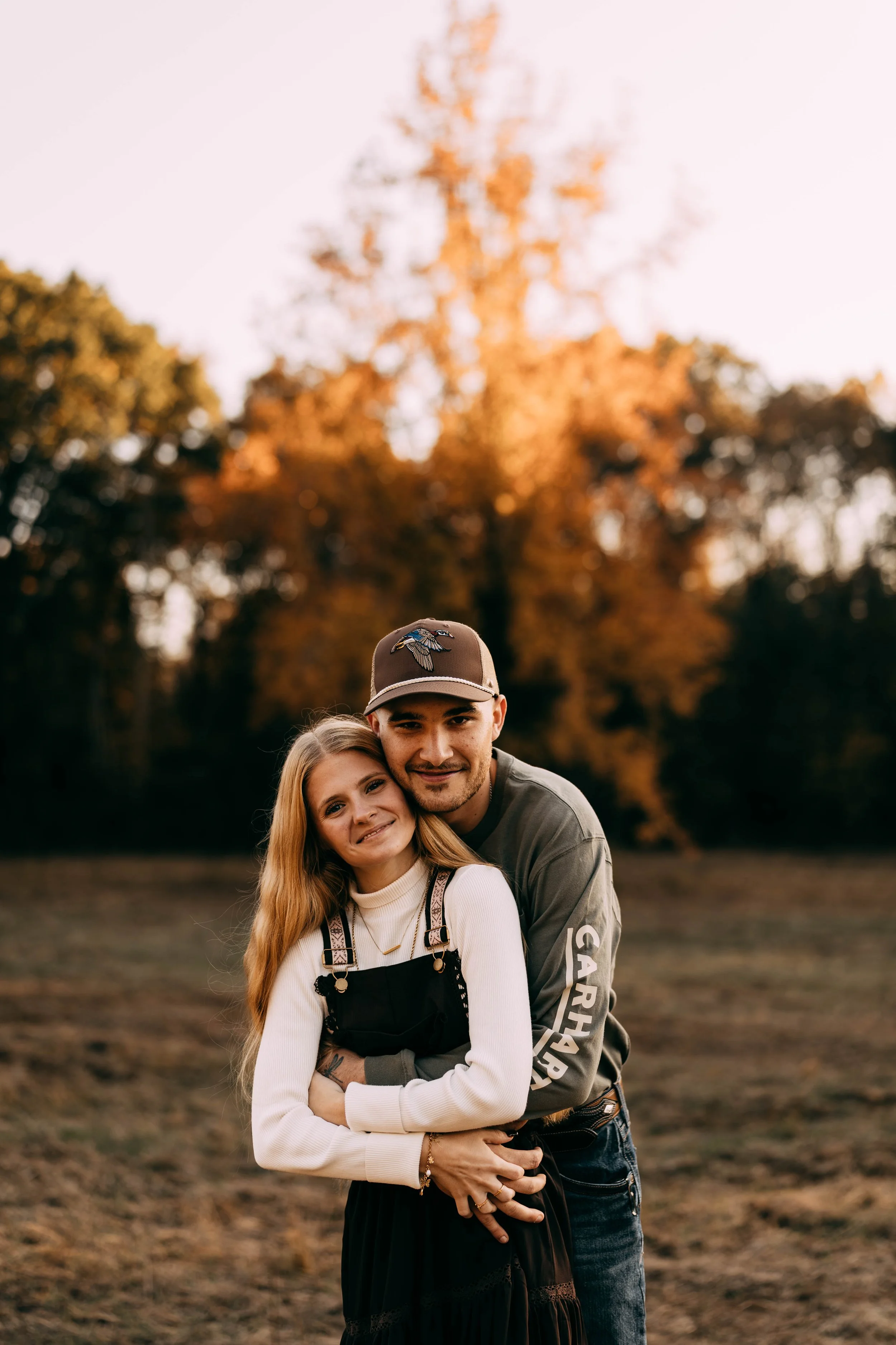 A young couple with light skin hugging outdoors on a fall evening, with trees showing autumn leaves in the background.