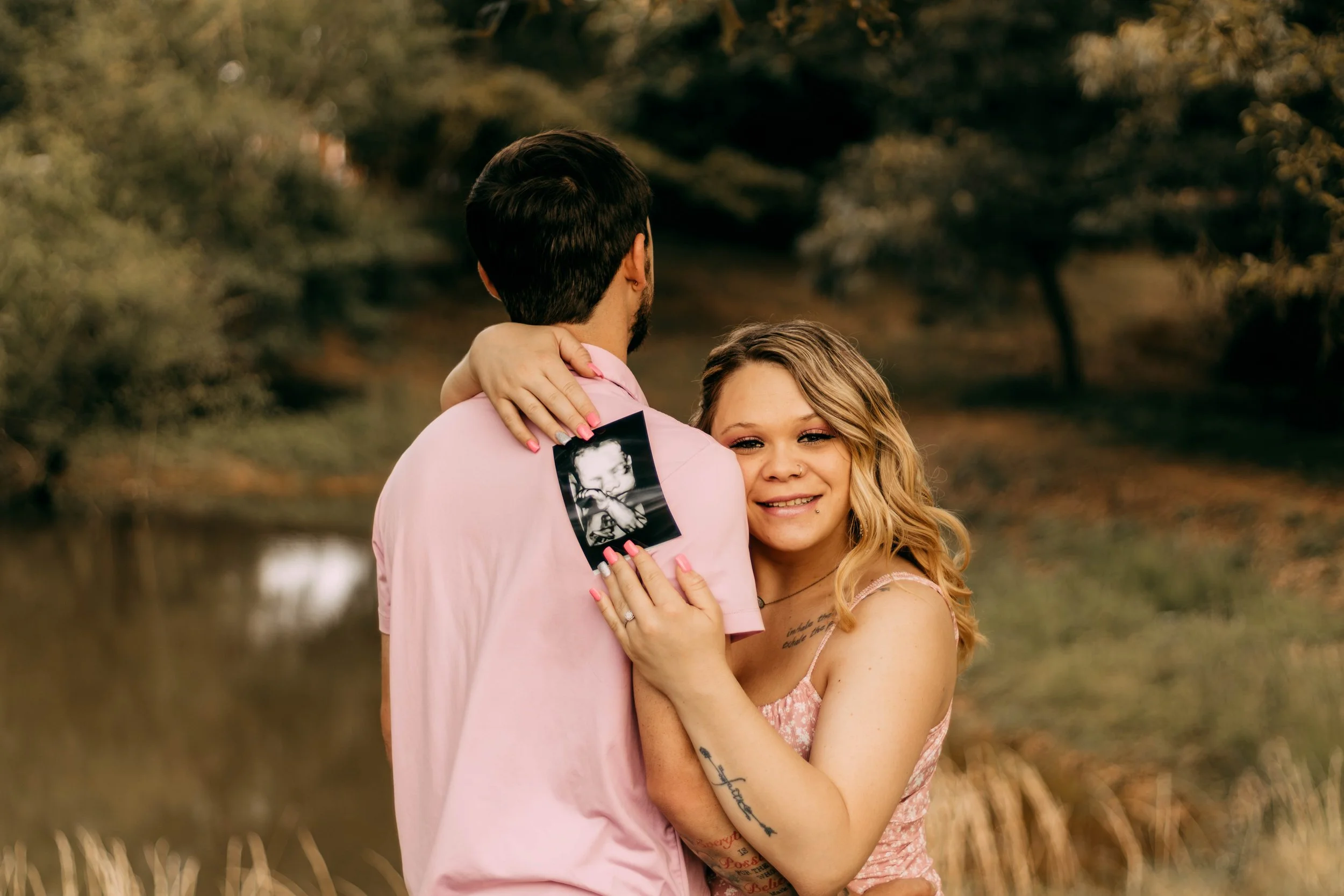 A blonde woman with tattoos on her arms and wearing a pink dress hugs a man with dark hair wearing a pink shirt. The woman is holding an ultrasound photo on the man's back while smiling at the camera. They are outdoors near a body of water with trees