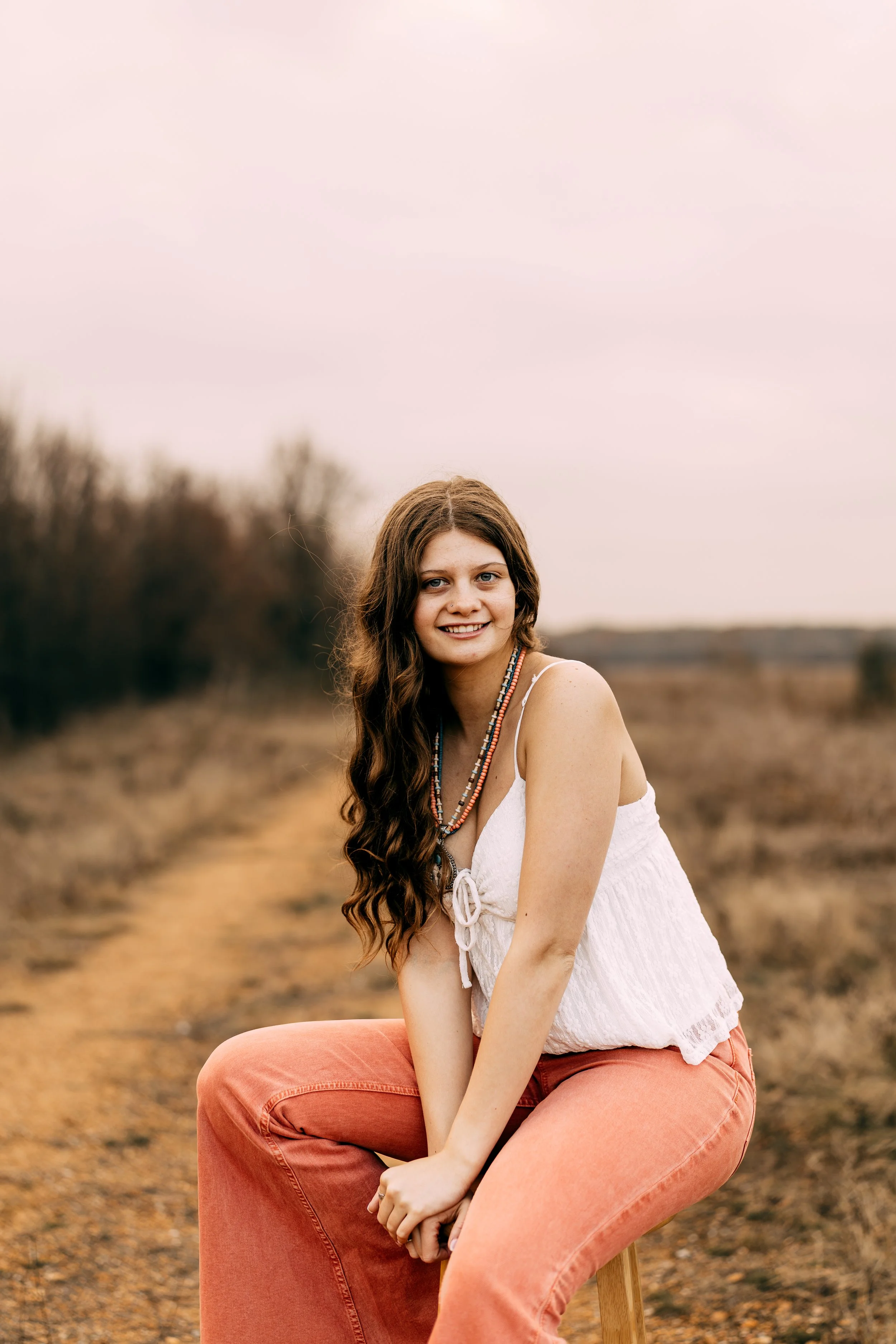 A young woman with long, wavy brown hair smiling while sitting on a wooden stool in an outdoor setting with a dirt path and trees in the background during dusk.