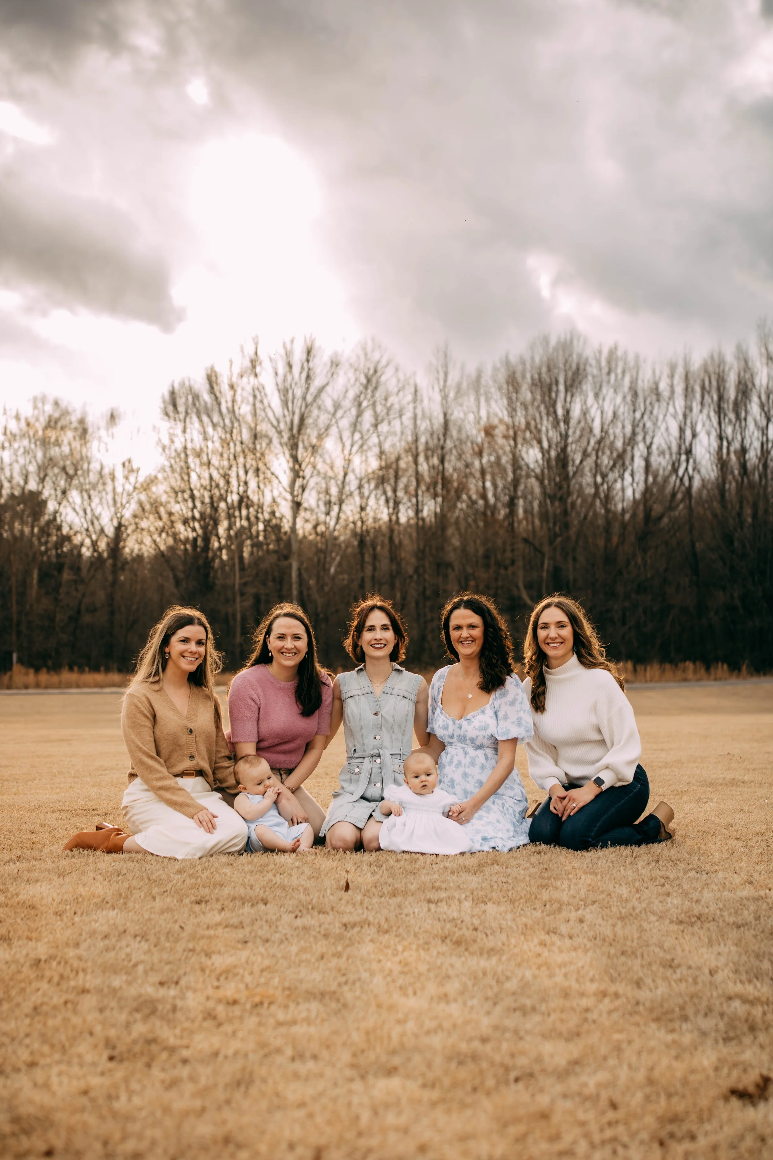 A group of five women and two babies sitting on grass outdoors, with a backdrop of leafless trees and an overcast sky.