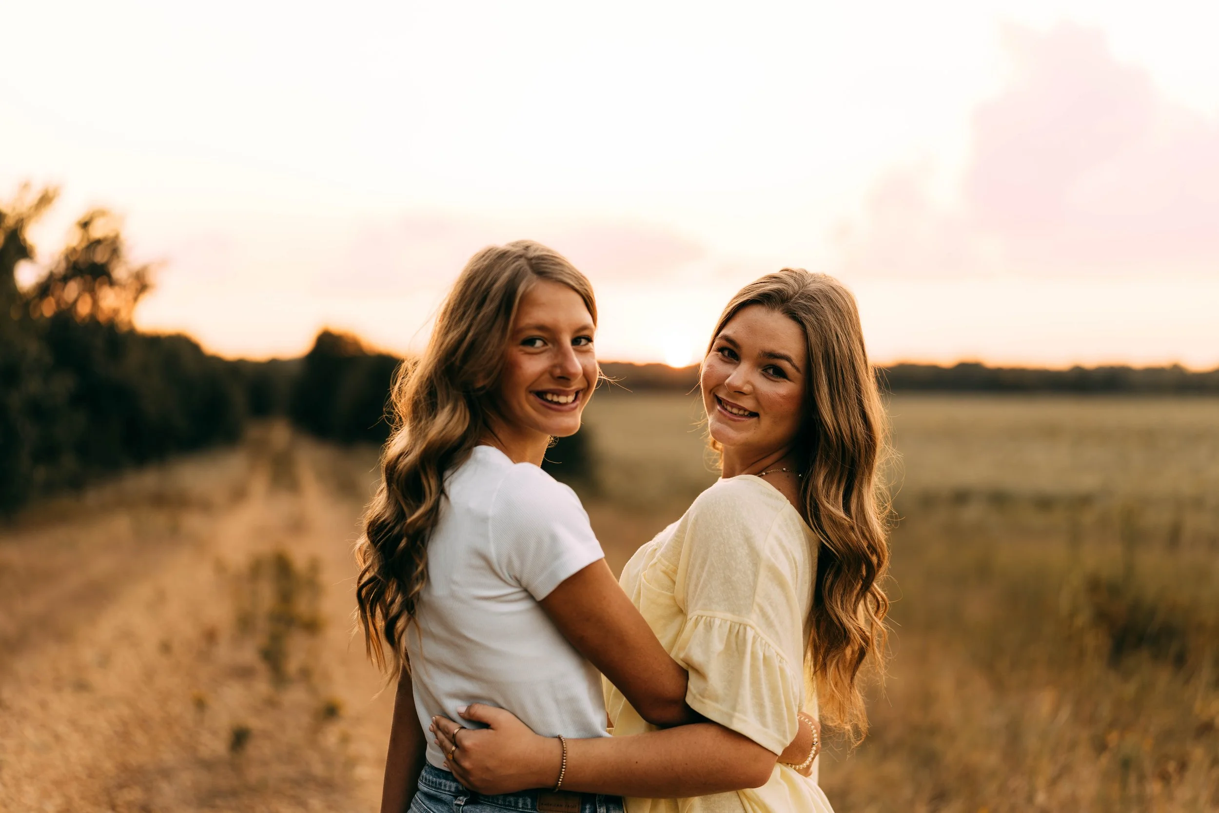 Two young women smiling and embracing outdoors during sunset in a field.