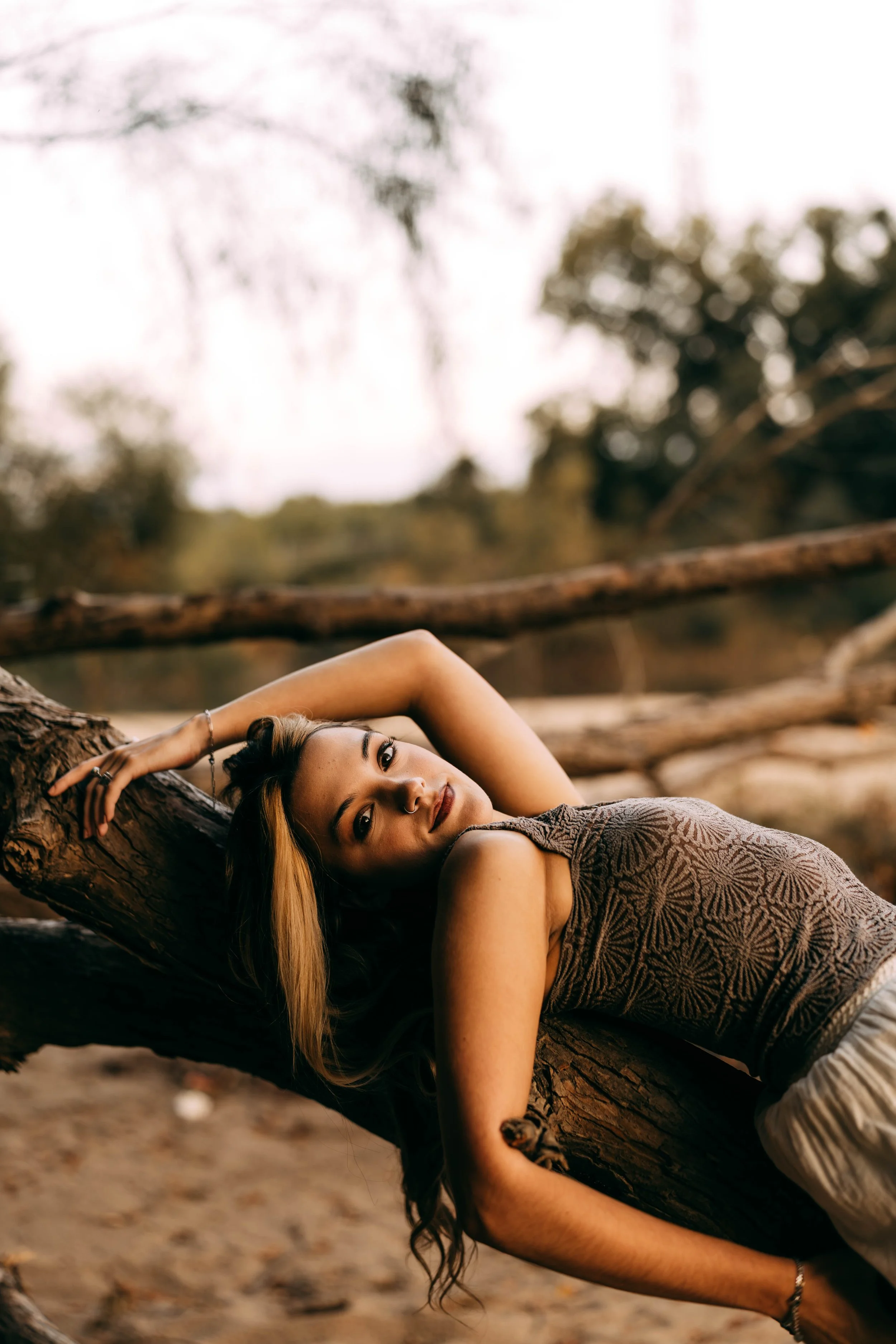 A woman with long hair lying on a fallen tree trunk outdoors in nature during sunset or sunrise, gazing at the camera.