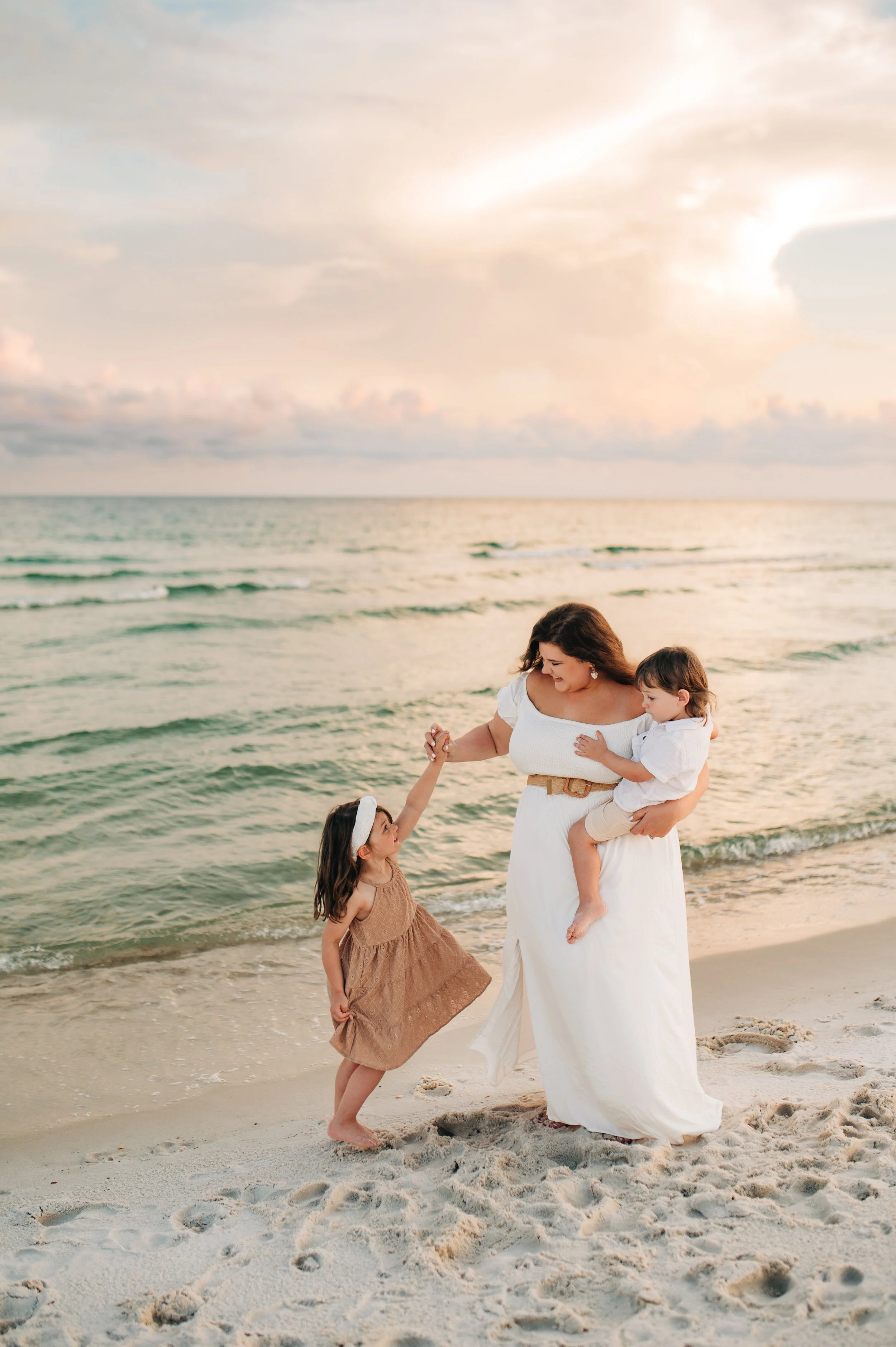 A woman holding a young boy in her arms on a sandy beach at sunset, dancing with a young girl who is looking up and holding her hand.
