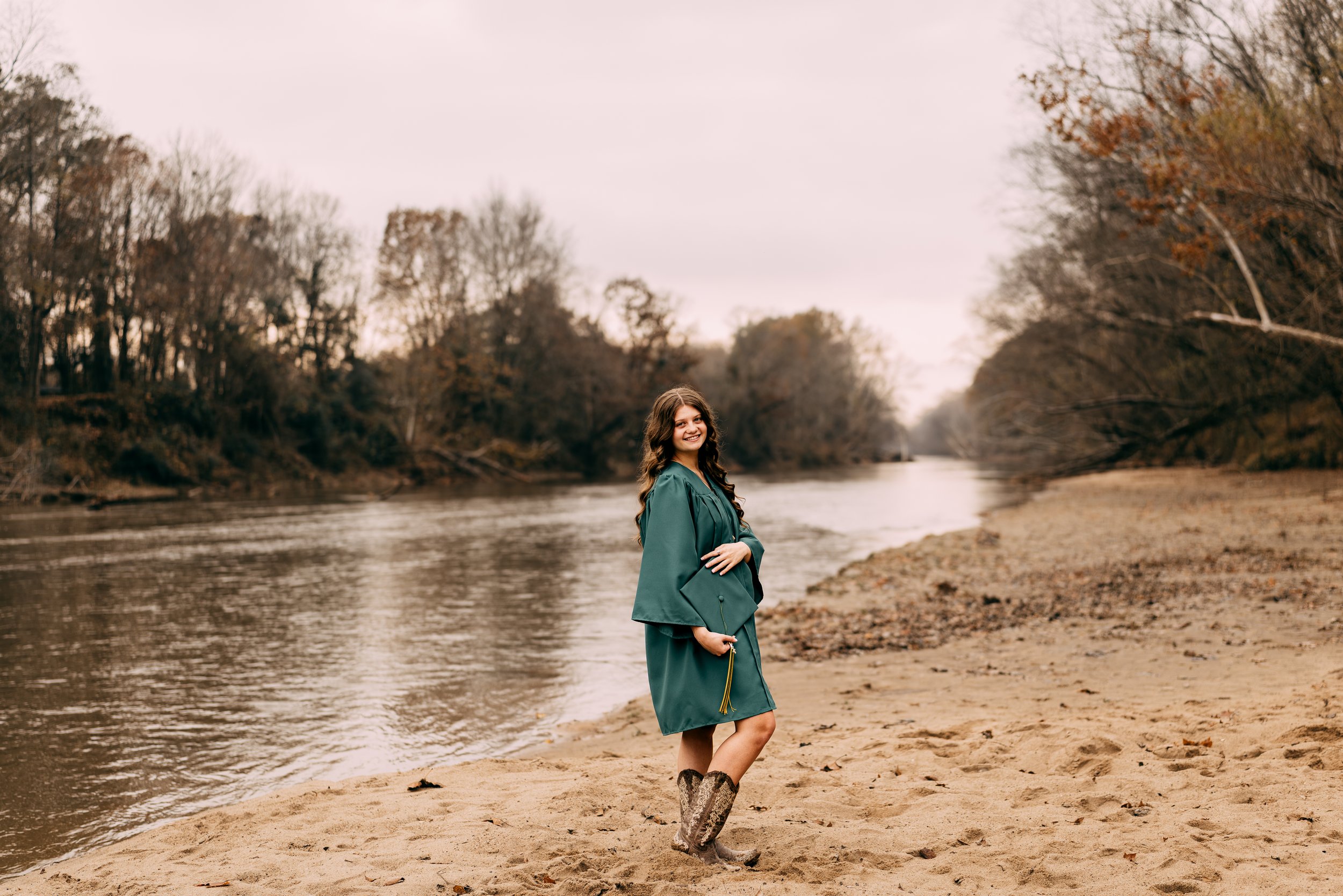 A young woman in a teal graduation gown holding a cap, standing on a sandy riverbank with trees and water in the background, smiling at the camera.