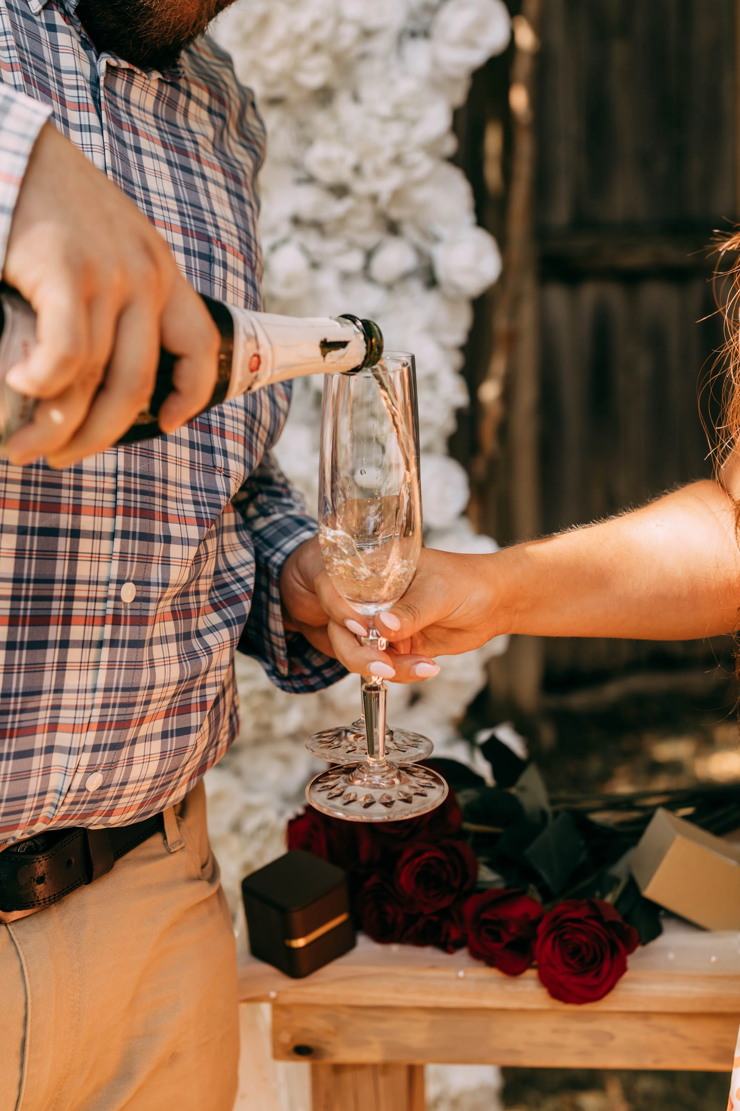 Person pouring champagne into a glass while another person holds the glass, with roses and a gift box on a wooden table in the background at a celebration or romantic setting.