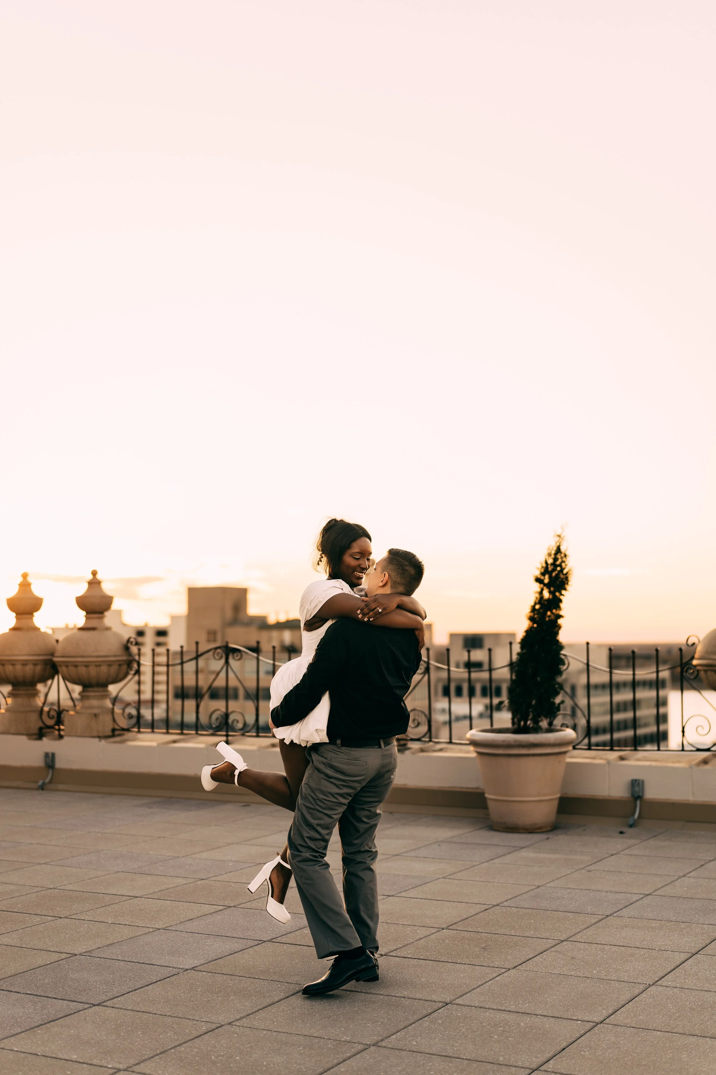 A couple dressed in formal attire embracing on a rooftop during sunset, with city buildings in the background.